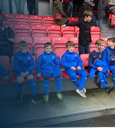 Four young boys in blue soccer uniforms sitting on a bench, with a few adults and other children standing behind them in a stadium.