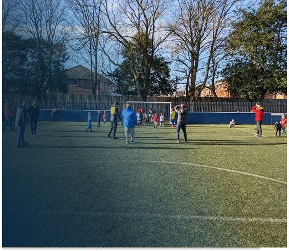 Children and adults are playing and walking on a grassy sports field enclosed by a fence, with tall trees and houses in the background on a clear day.