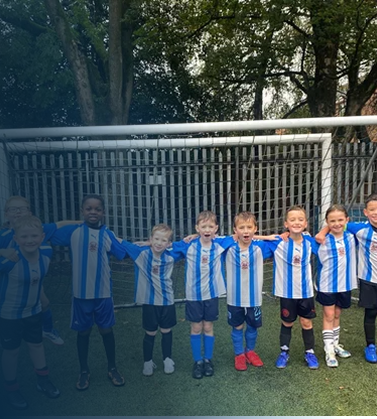 Group of young boys in soccer uniforms standing in a line on a soccer field, smiling, with a metal fence and trees in the background.