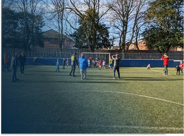 Kids and adults on a soccer field, some preparing to play or learn, with a fence, trees, and houses in the background on a sunny day.