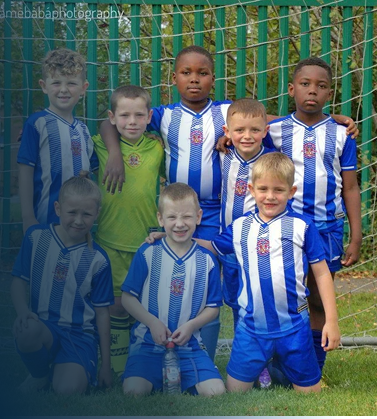 Group of seven young boys in blue and white soccer jerseys posing for a photo outdoors, with a green fence in the background.