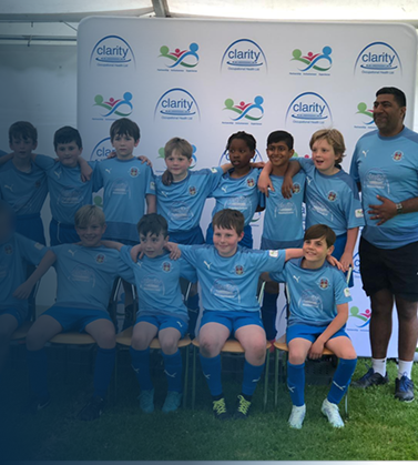 Youth soccer team in light blue jerseys posing with their coach in front of a Clarity sponsorship banner.