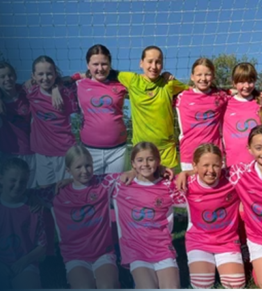 Group of young female soccer players in pink jerseys and white shorts, standing together outdoors after a game, smiling.