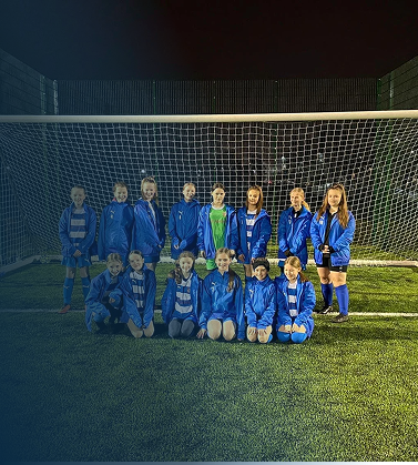 A girls' soccer team in blue uniforms posing in front of a goal on a soccer field at night.