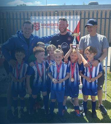 Young boys in blue and white soccer uniforms with medals around their necks, posing with three adult men outdoors on a sunny day, one boy holding a small trophy, in front of a fence and a banner that says 'ACCREDITED'.
