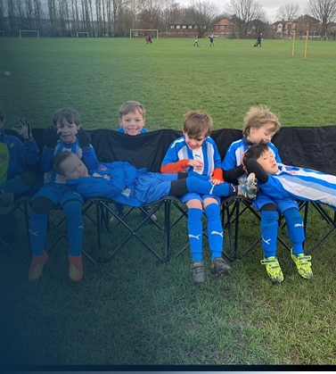Young boys in blue soccer uniforms sitting on a bench during a game on a grassy field.