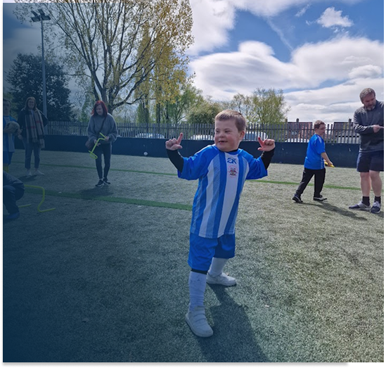 Child in a blue and white sports jersey and shorts standing on a field, smiling and raising hands, with other children and adults in the background under a partly cloudy sky.