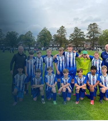 Youth soccer team in blue and white jerseys posing on a field with coaches, with trees and buildings in the background.