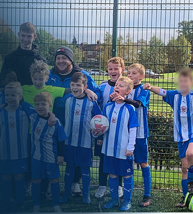 Group of young boys and two adults, one holding a soccer ball, on a soccer field with a fence and trees in the background