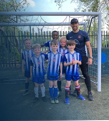 A group of young boys and an adult male coach posing in front of a soccer goal. The boys are wearing blue and white striped soccer jerseys and shorts, and the coach is dressed in a navy blue shirt and black pants. They are outdoors on a paved soccer field with a wooden fence and trees in the background.