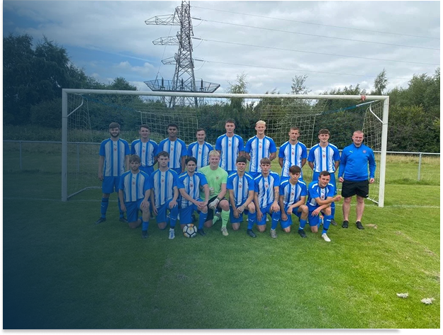 Group of young soccer players in blue and white uniforms with two coaches, posing on a soccer field in front of a goal post, with power lines and trees in the background.