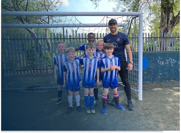 Group of six young soccer players and their coach standing in front of a soccer goal on a field, all smiling.