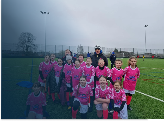 Youth girls' soccer team in pink jerseys and black shorts on a field, posing with coaches on a cloudy day.