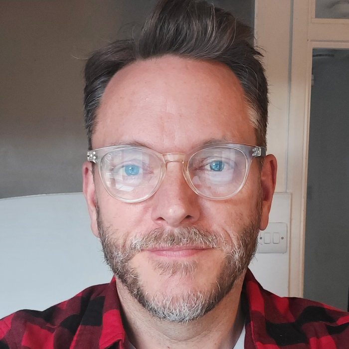 A man called Roly smiling wearing glasses and a red checked shirt.