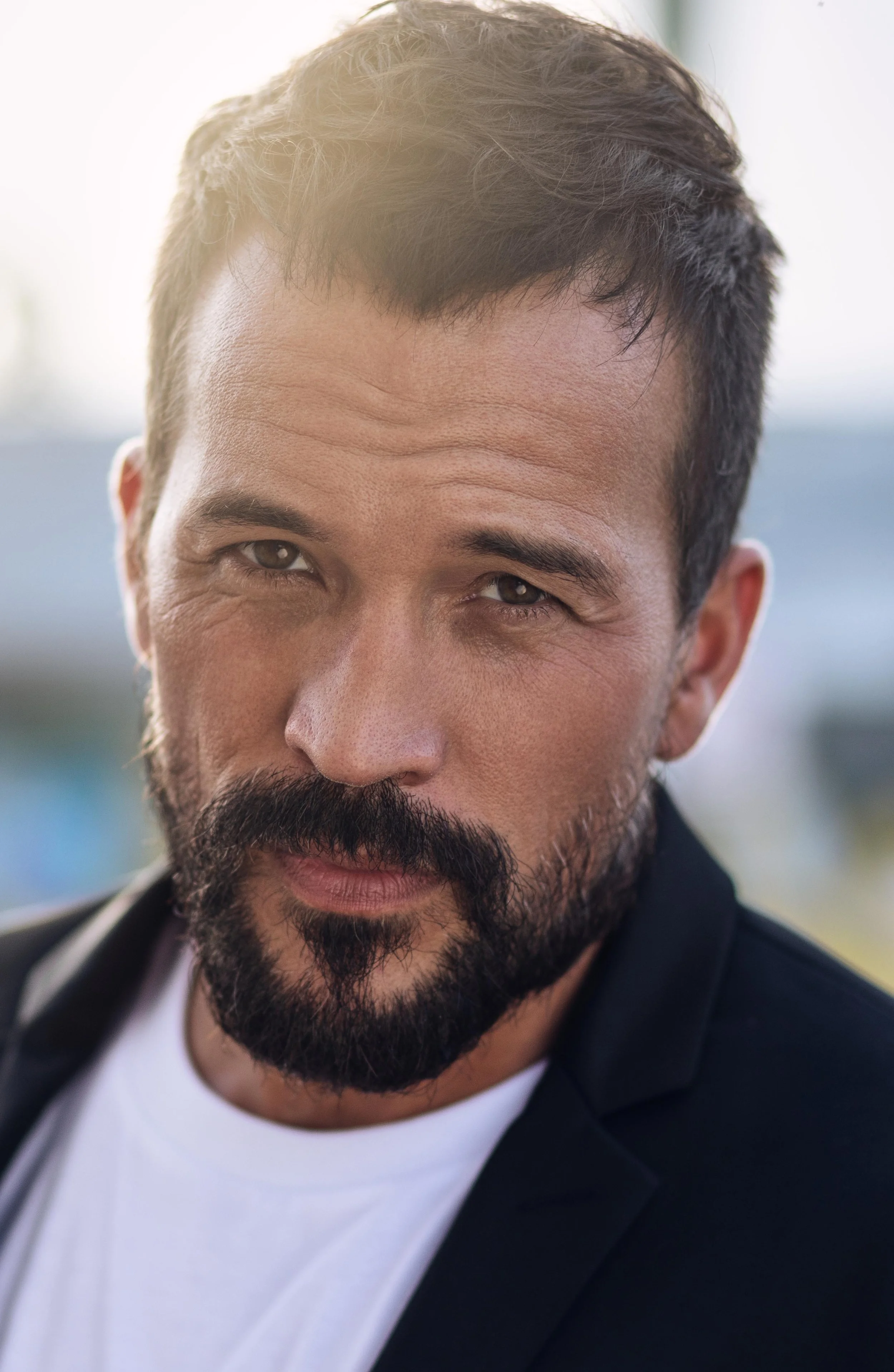 Close-up of a man with dark hair, a beard, and mustache, looking into the camera with a slight smile. He is wearing a dark jacket over a white shirt, with a blurred outdoor background and sunlight.