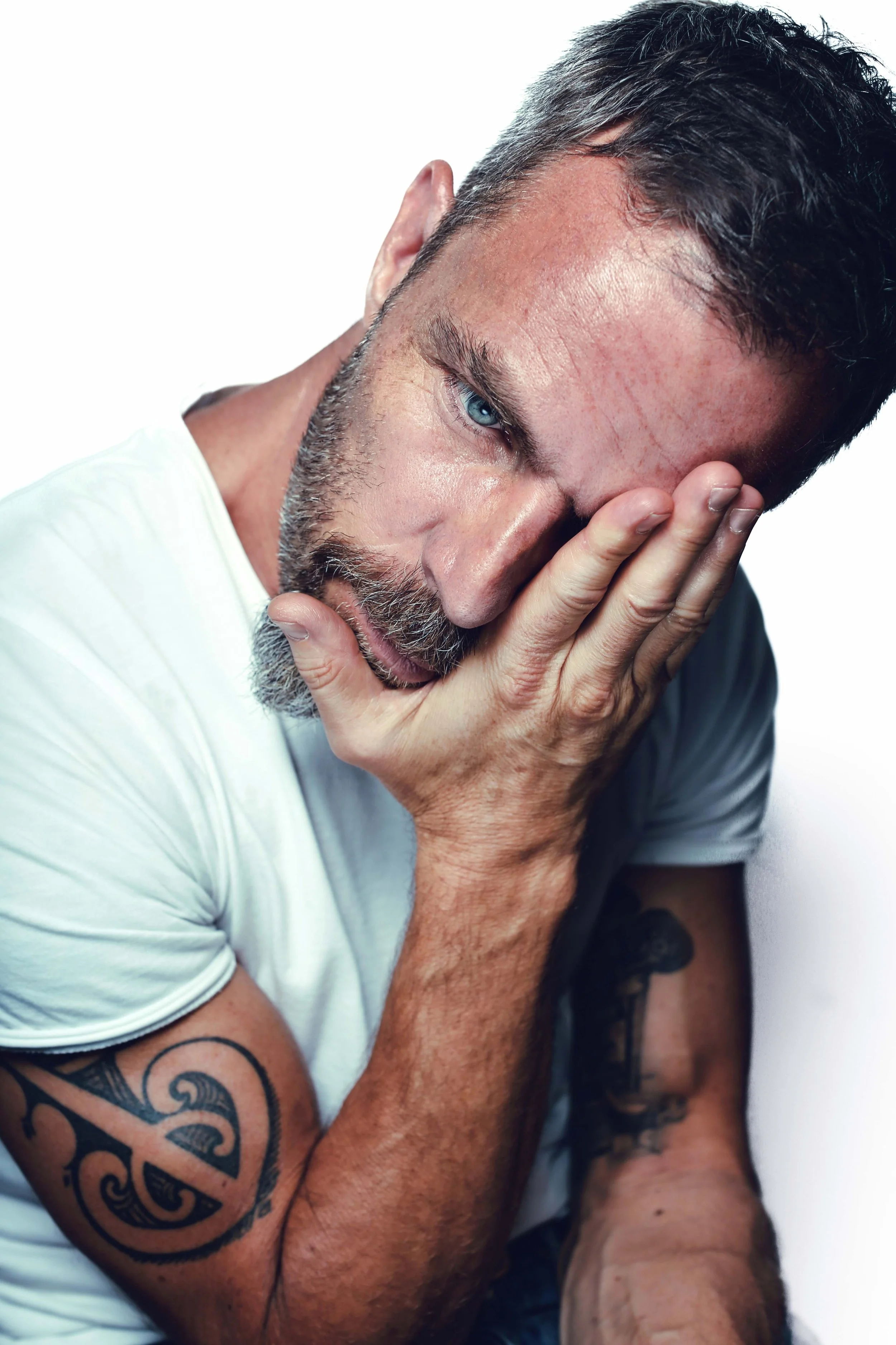 Close-up of a man with short dark hair, blue eyes, and tattoos on his arms, wearing a white shirt. He is covering part of his face with his hand, looking distressed against a plain white background.
