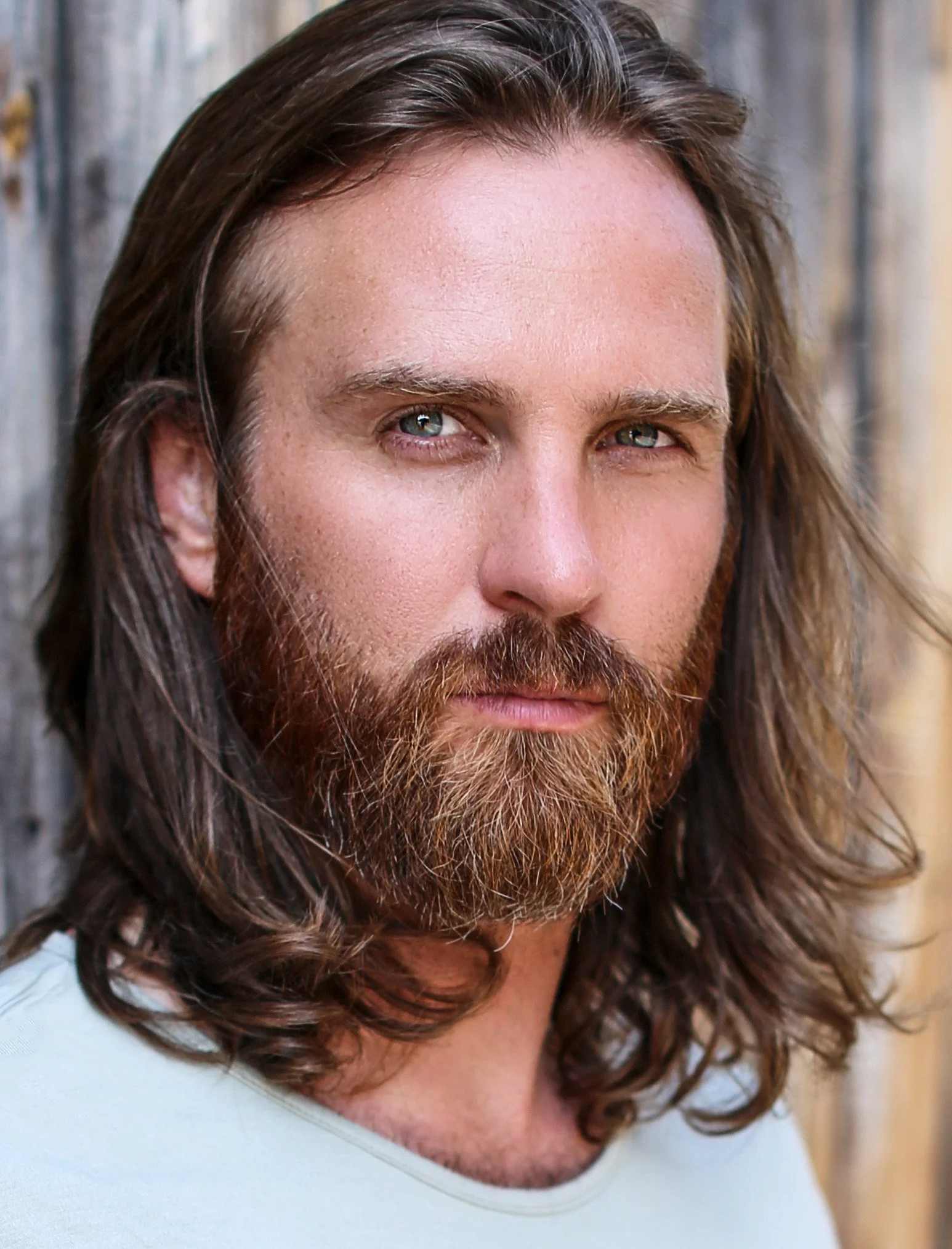 Close-up of a man with long brown hair, blue eyes, and a full beard, standing next to a wooden wall.