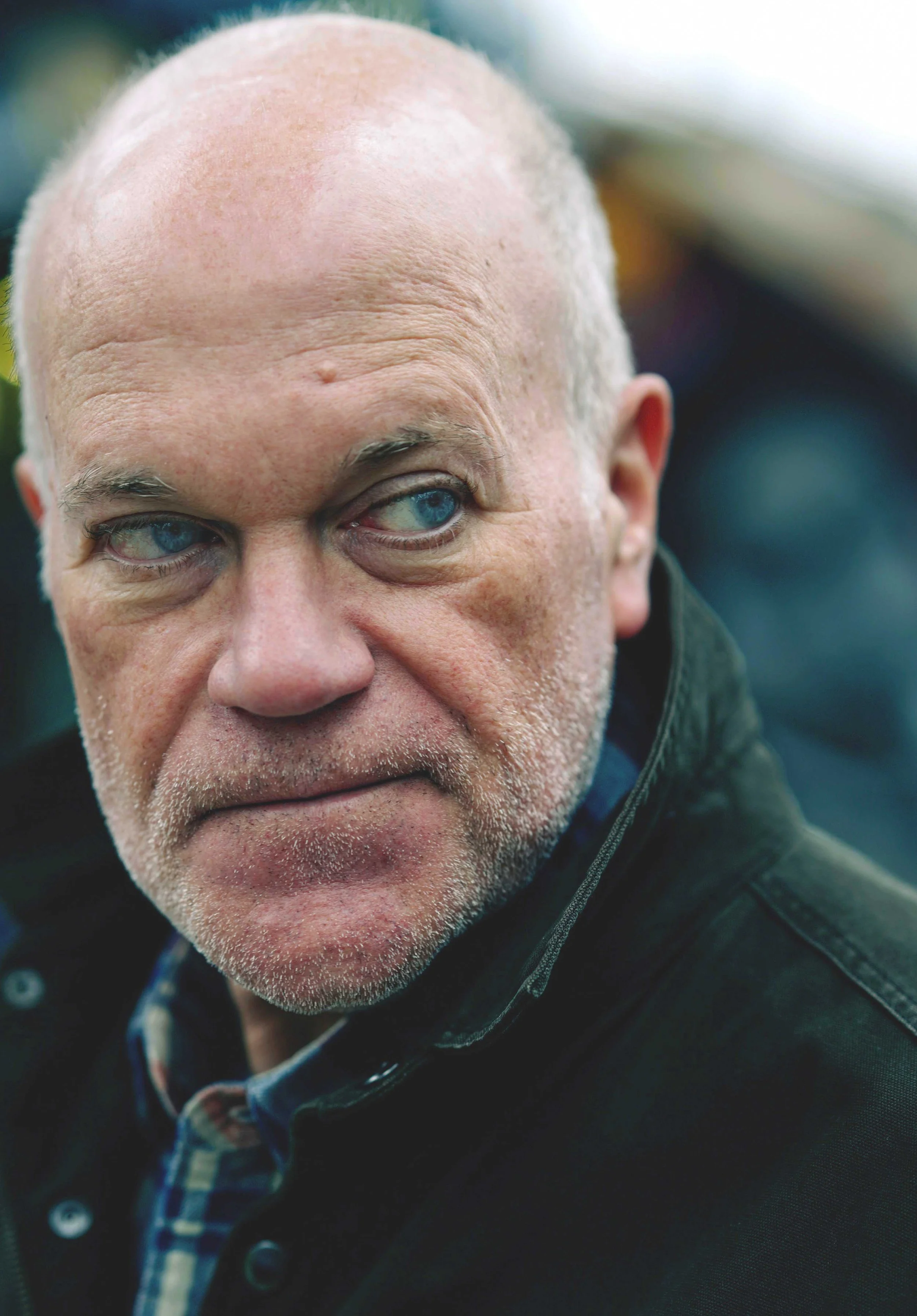 Close-up of an older man with a bald head, blue eyes, and stubble, looking to the side, wearing a dark jacket and plaid shirt.