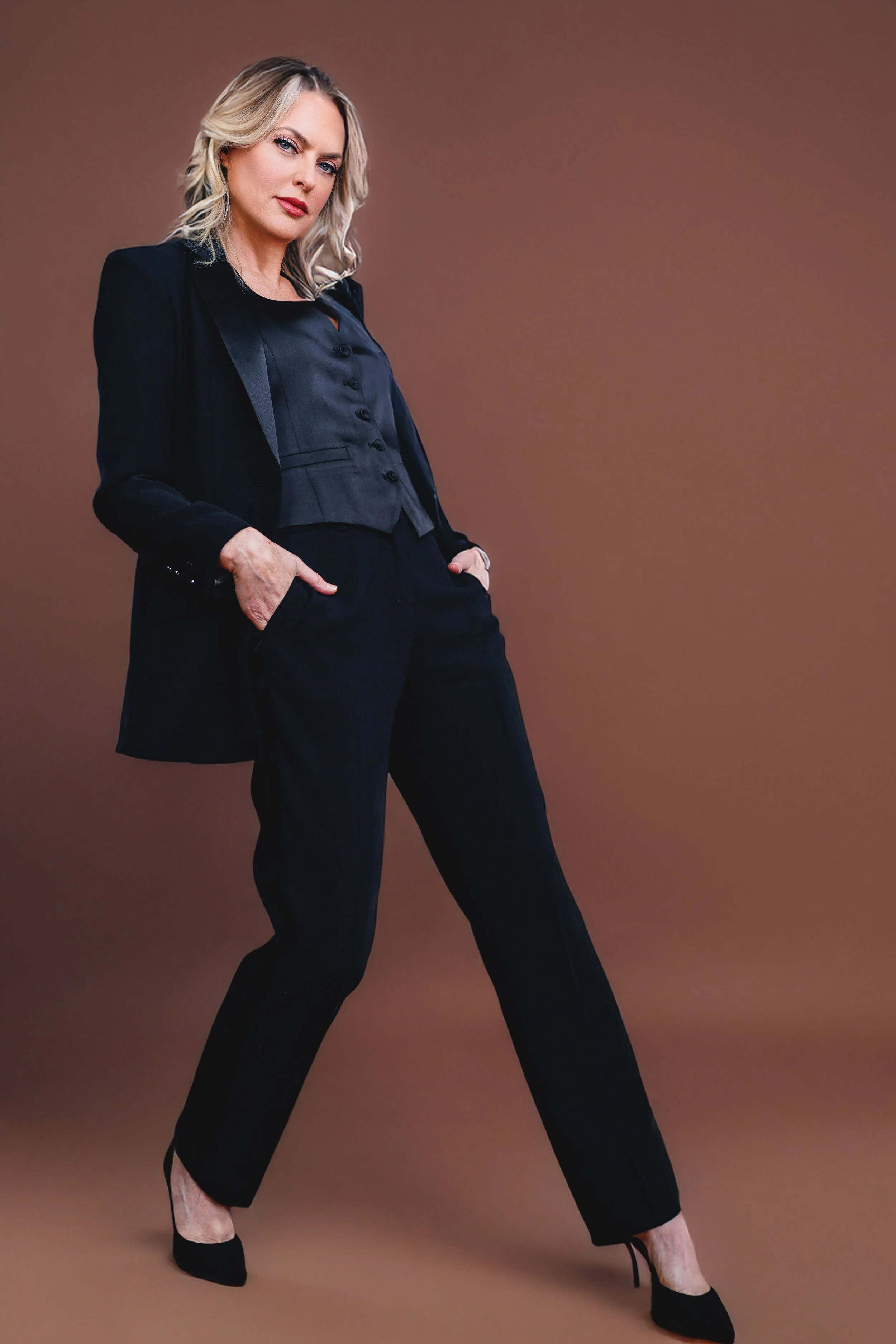 A woman in a black business suit standing with her hands in her pockets against a brown background.