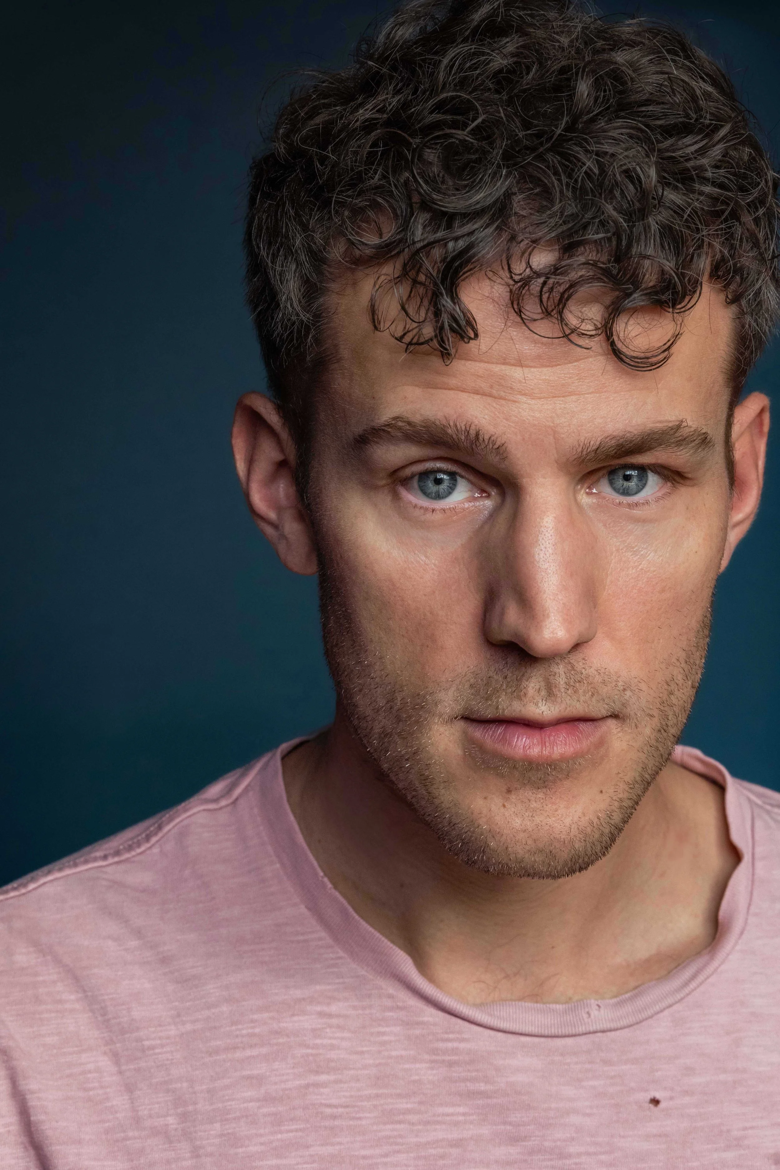 A close-up portrait of a young man with curly brown hair, blue eyes, and light skin, wearing a light pink T-shirt against a dark blue background.