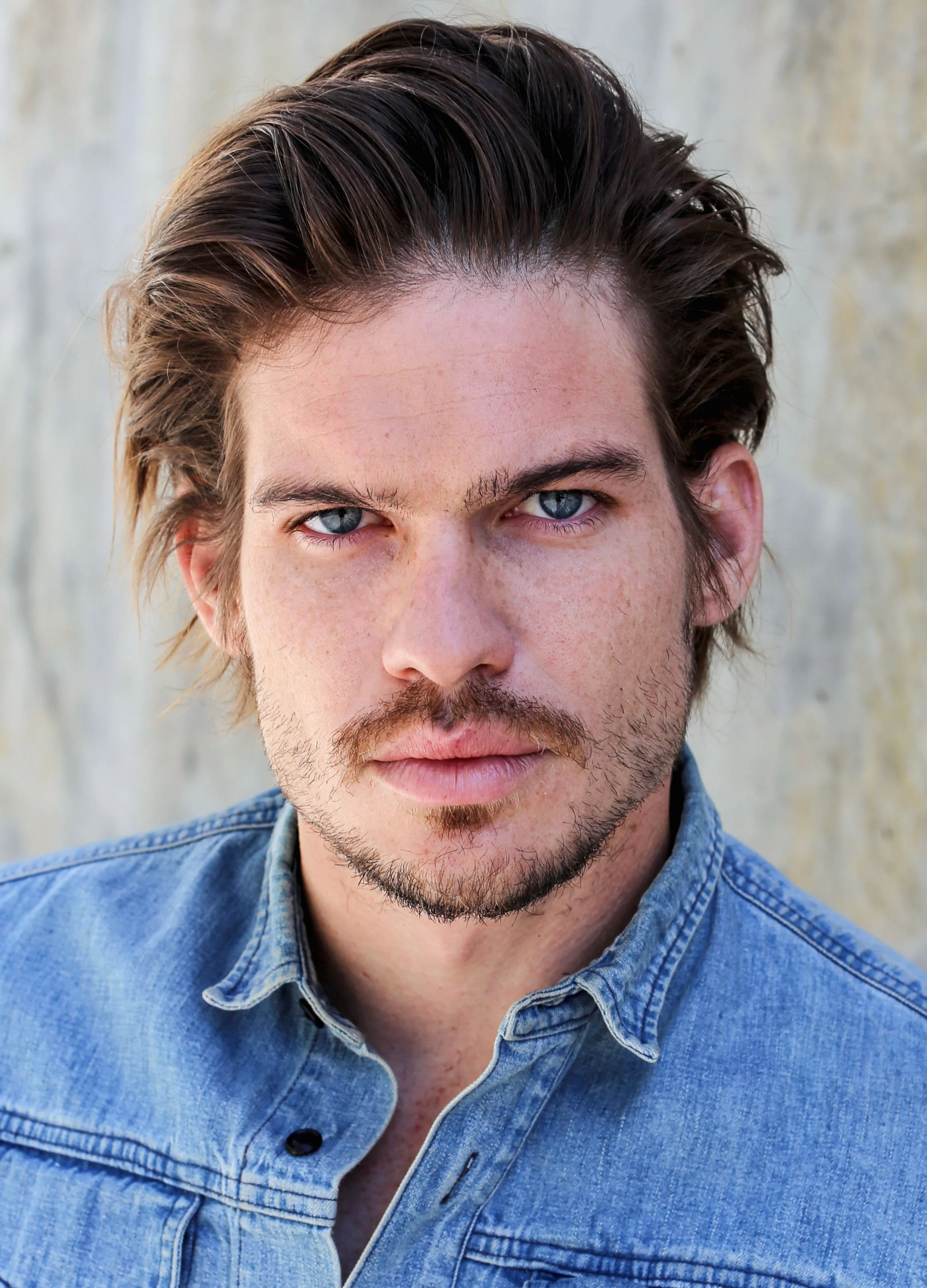 Close-up of a man with blue eyes, brown hair, and facial hair, wearing a denim shirt against a neutral background.