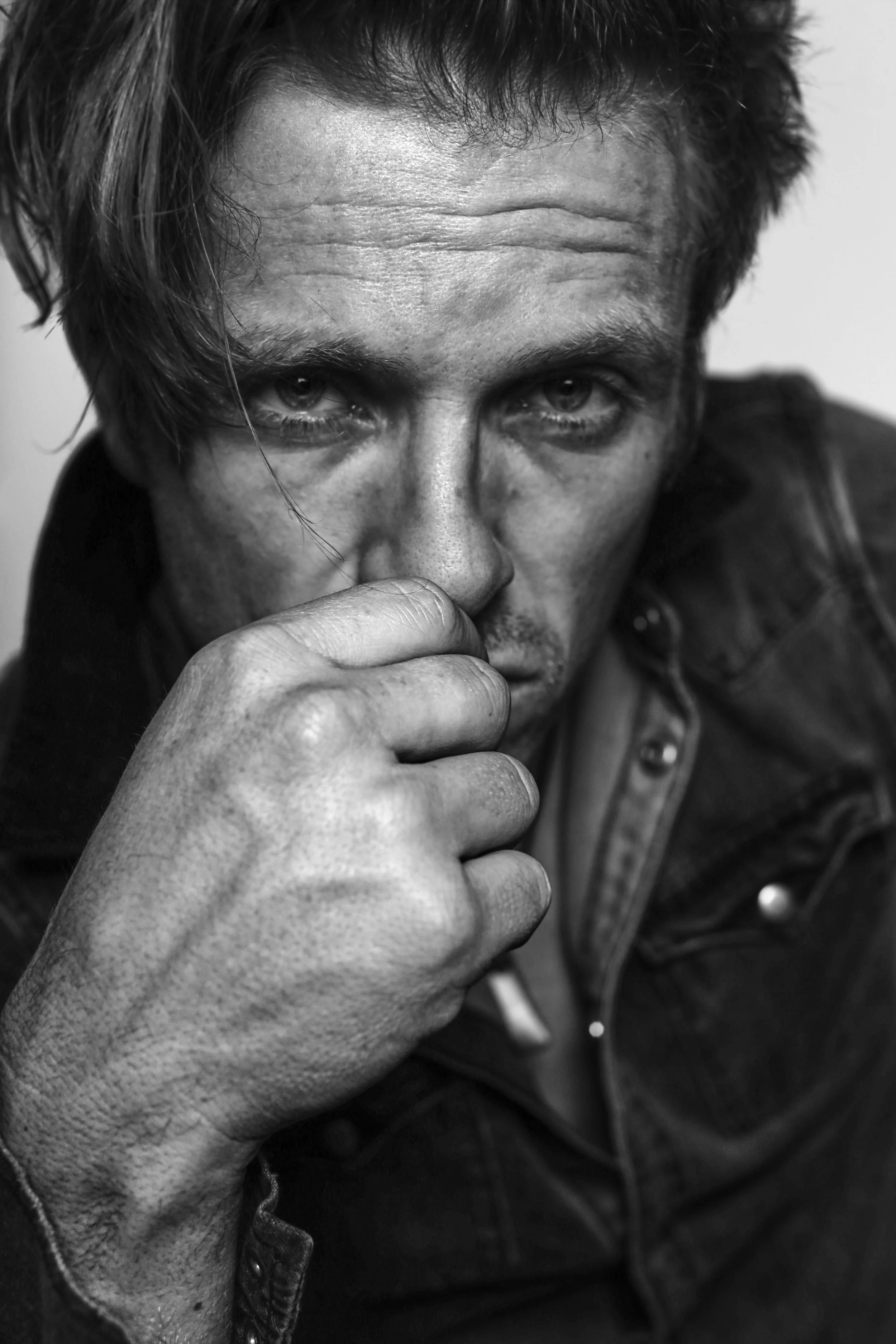 A black and white close-up portrait of a man with intense eyes, with some hair falling over his face, wearing a dark leather jacket, and resting his chin on his hand.
