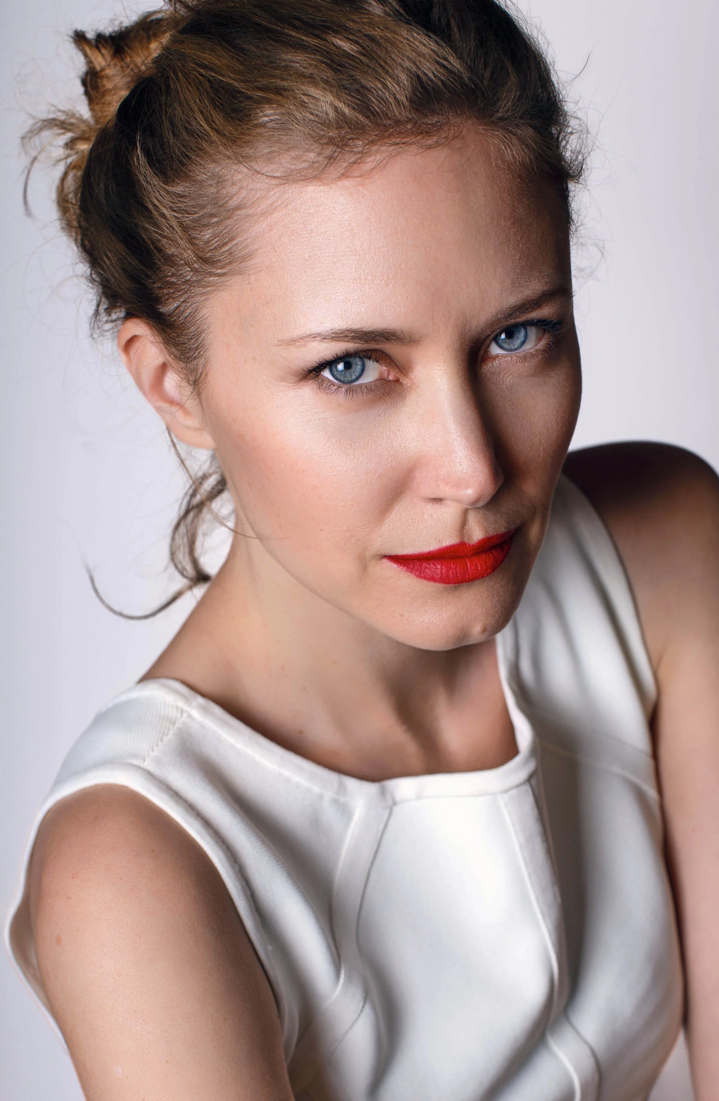 A woman with red lipstick and blue eyes wearing a sleeveless white top, looking at the camera.