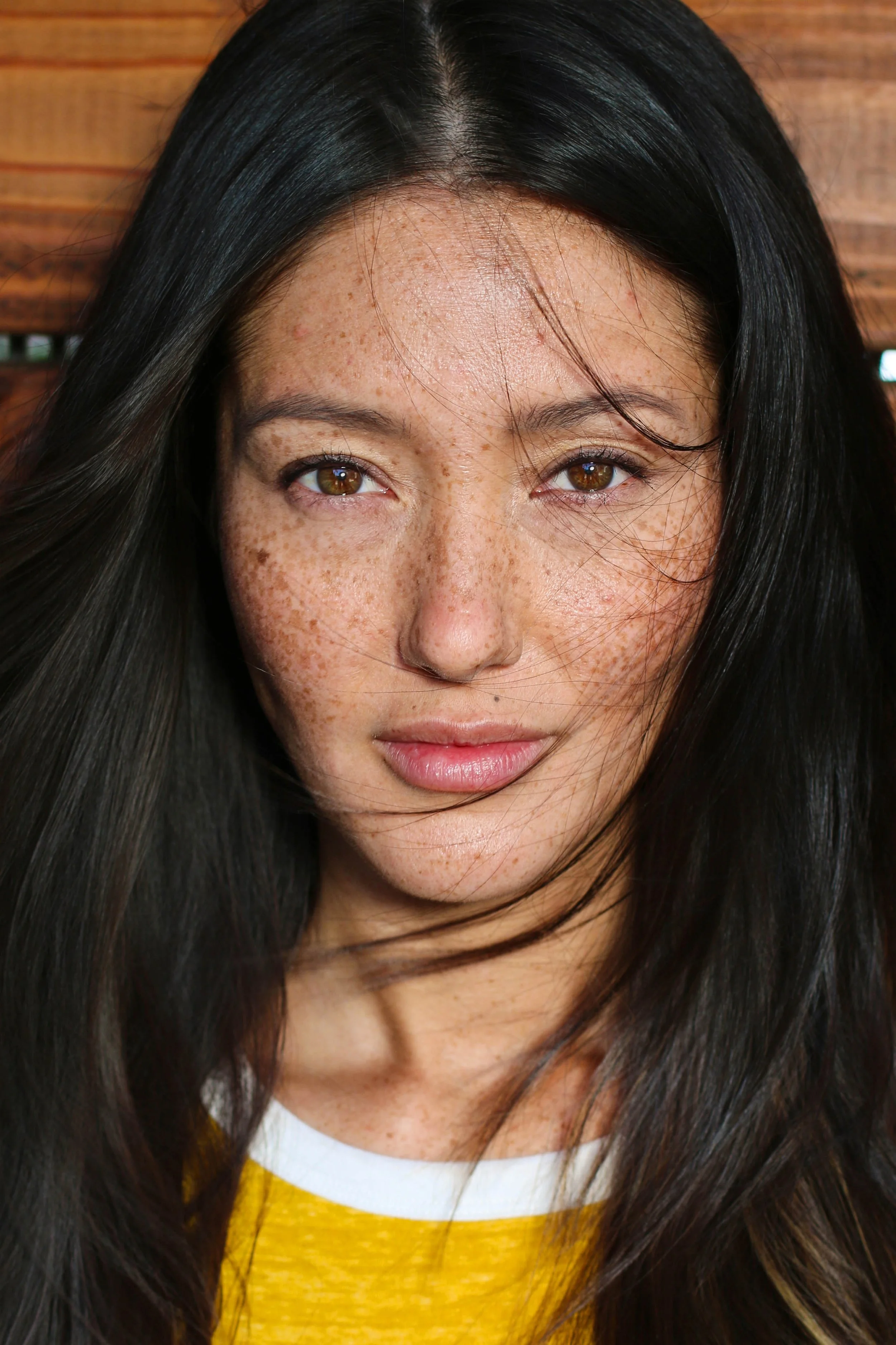 Close-up of a young woman with long black hair, brown eyes, and freckles, looking into the camera against a wooden background.