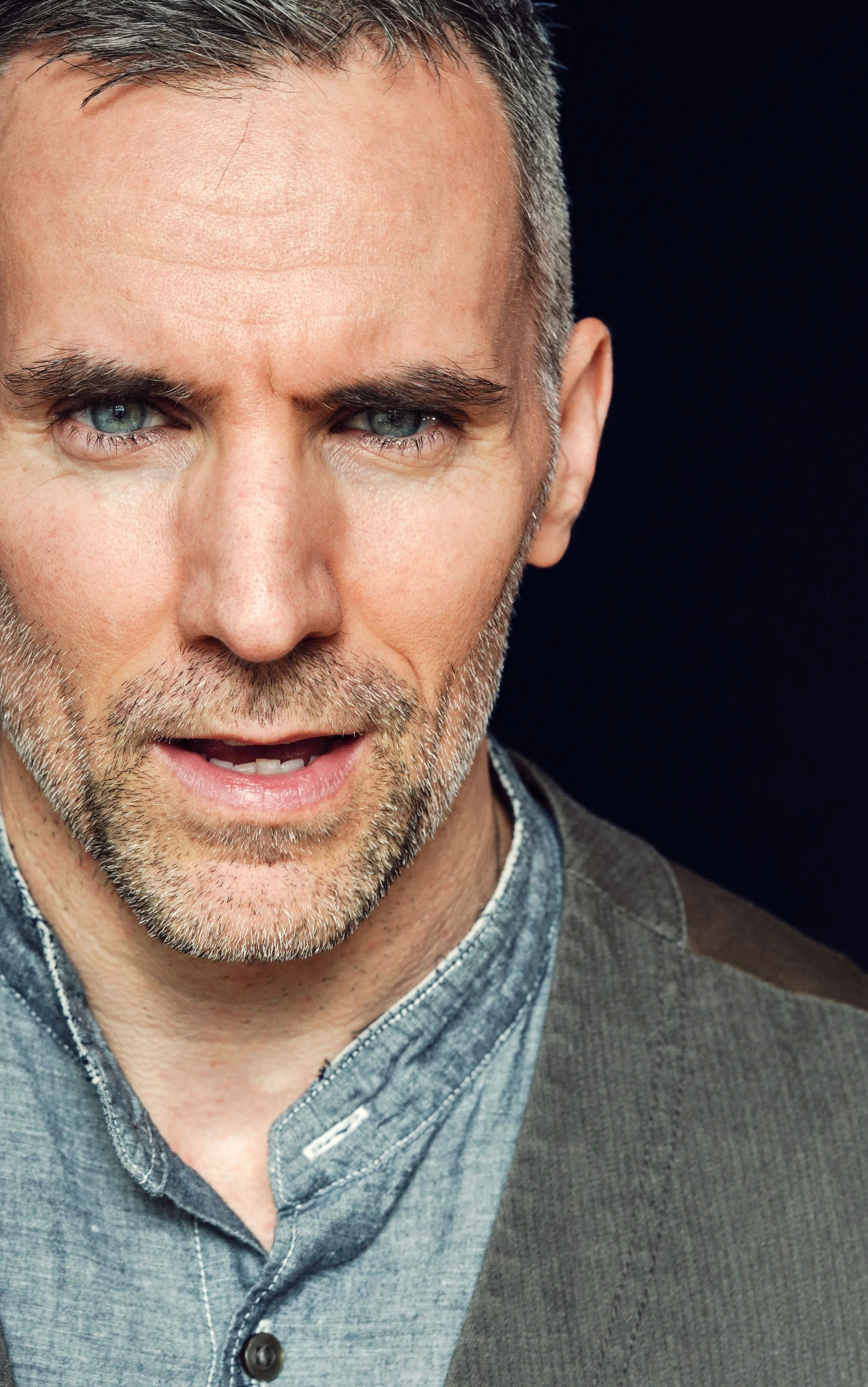 Close-up of a middle-aged man with gray hair and beard, wearing a gray shirt with a dark vest, against a black background.