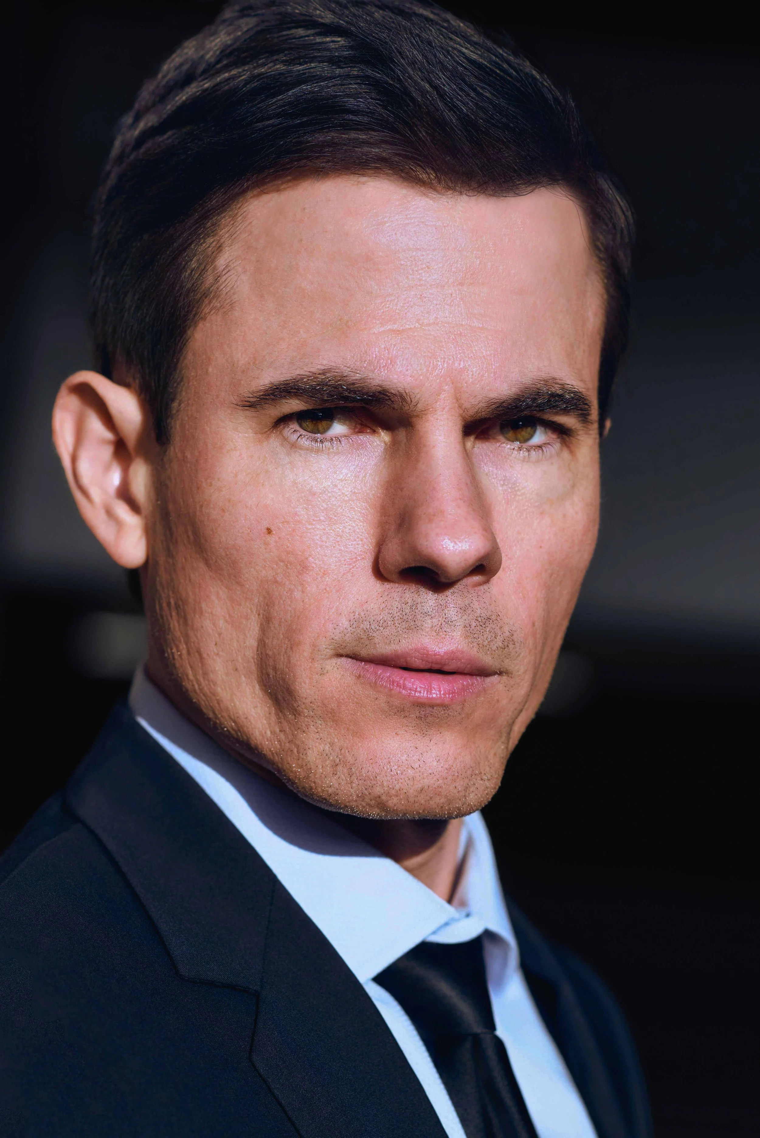 Close-up of a man with dark hair, wearing a suit and tie, against a dark background.