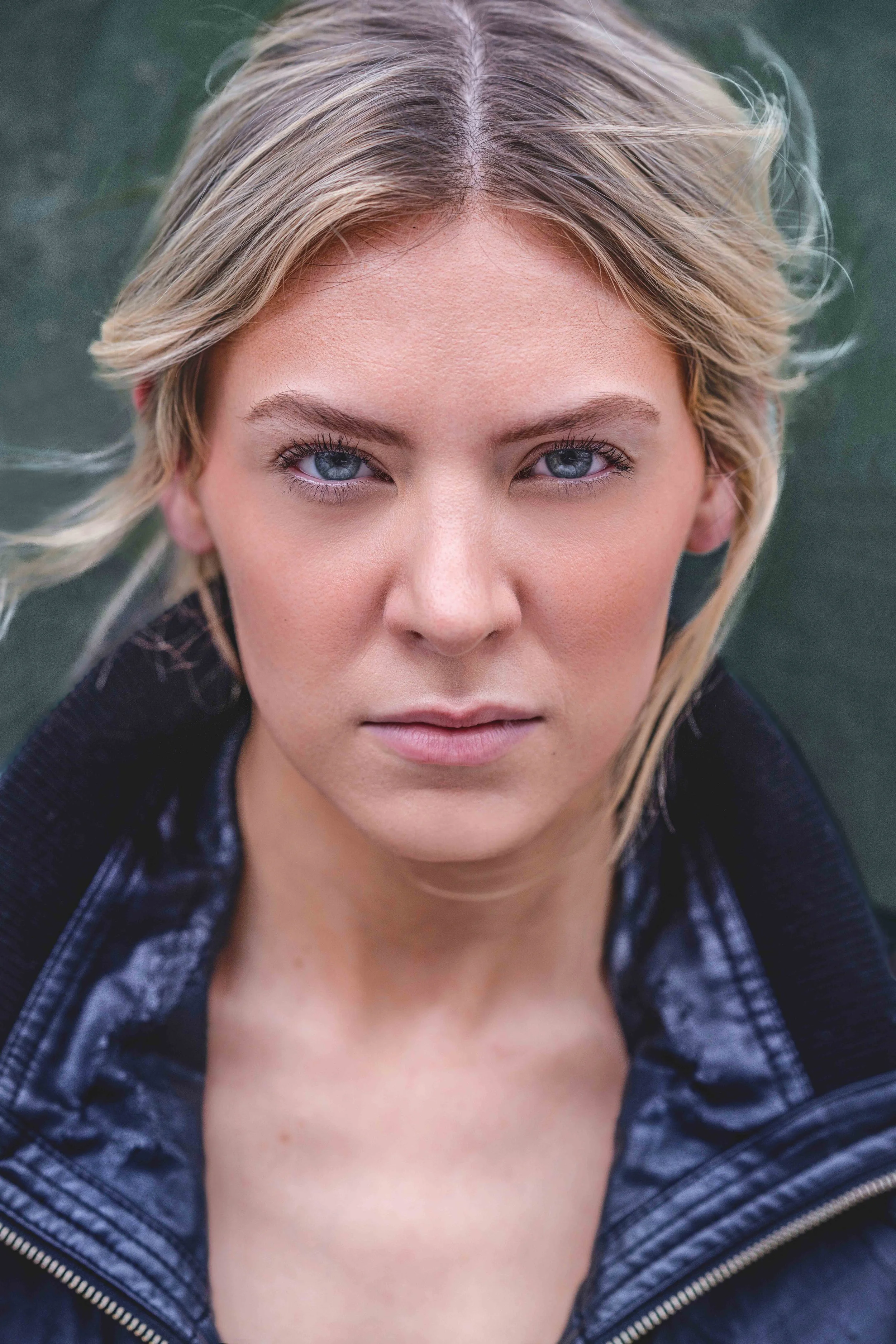 Close-up portrait of a young woman with blonde hair and blue eyes, wearing a black jacket, outdoors.