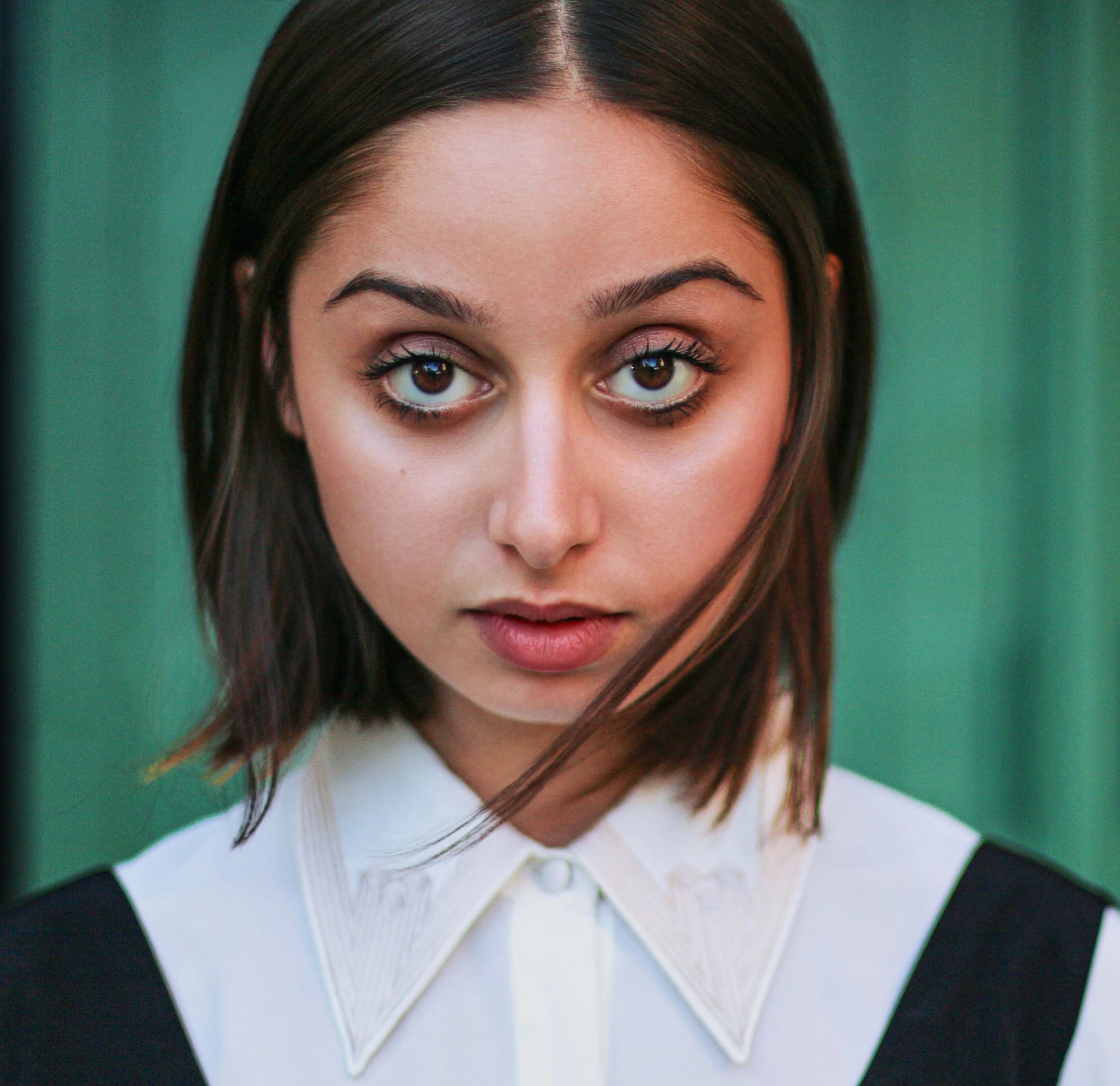 A young woman with shoulder-length brown hair wearing a white collared shirt with black accents, looking directly at the camera with a neutral expression against a teal background.