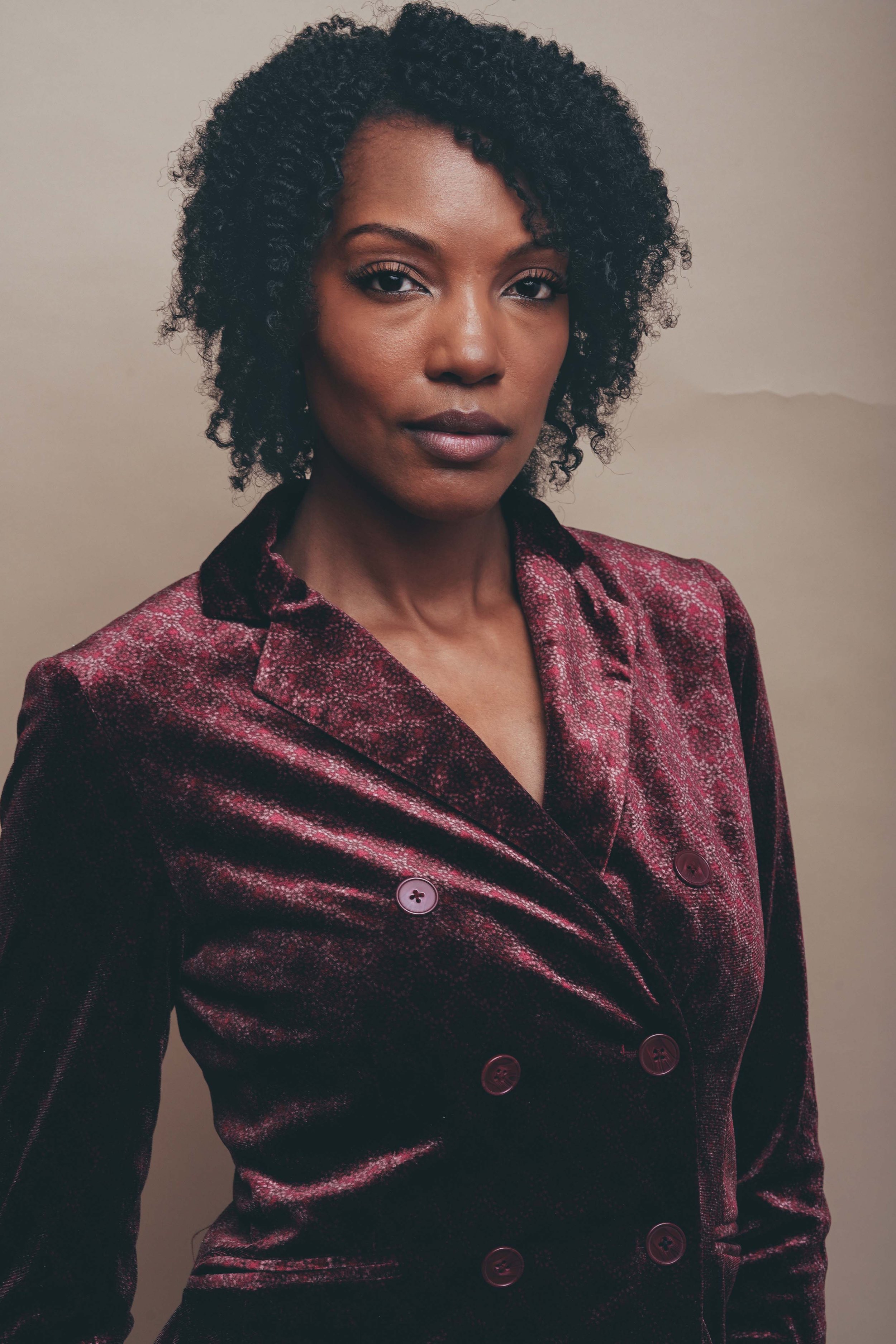 A woman with dark curly hair wearing a maroon velvet blazer with a subtle pattern, standing against a plain beige wall.