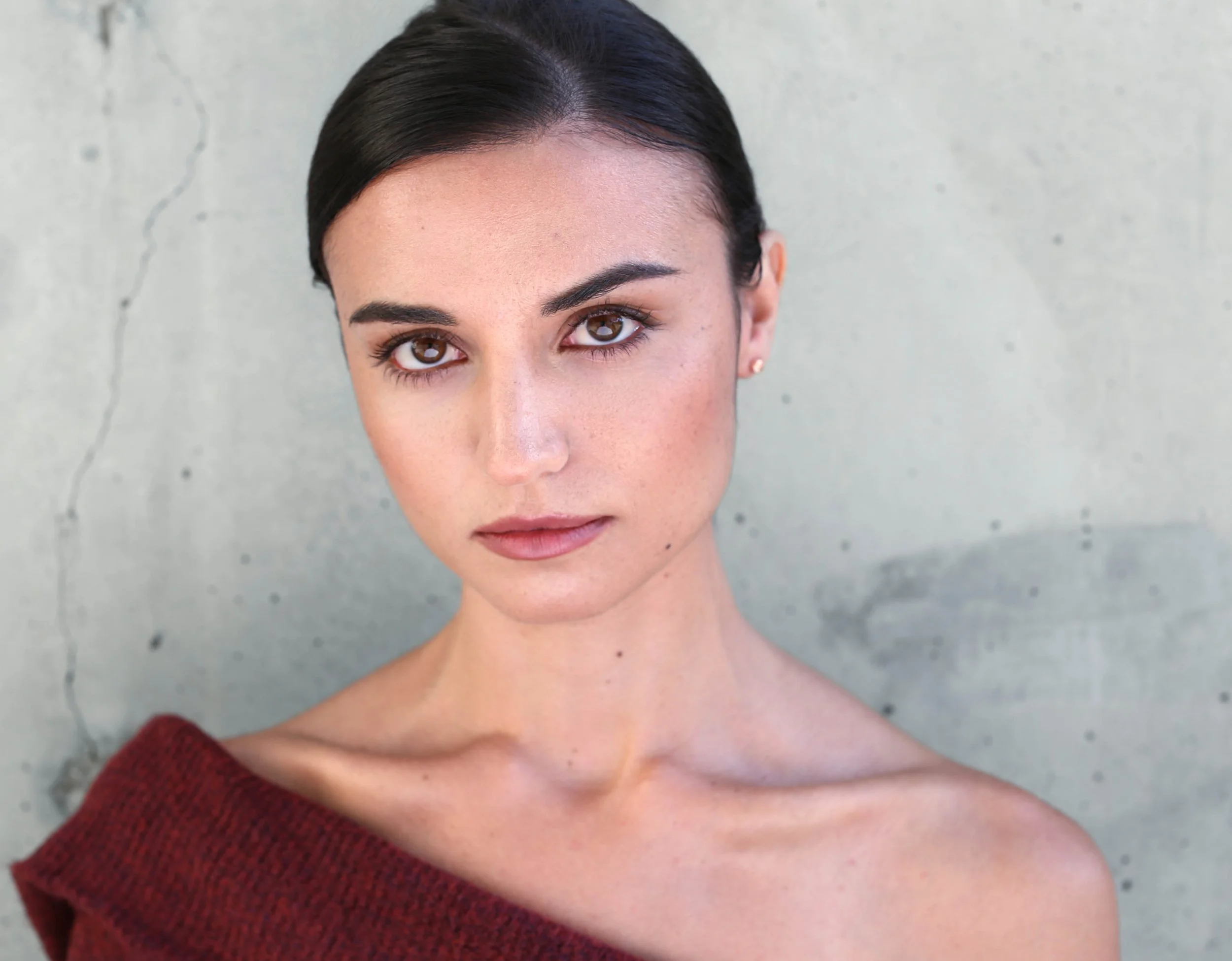 Close-up of a woman with short dark hair and brown eyes, wearing a red off-shoulder top, standing against a concrete wall.