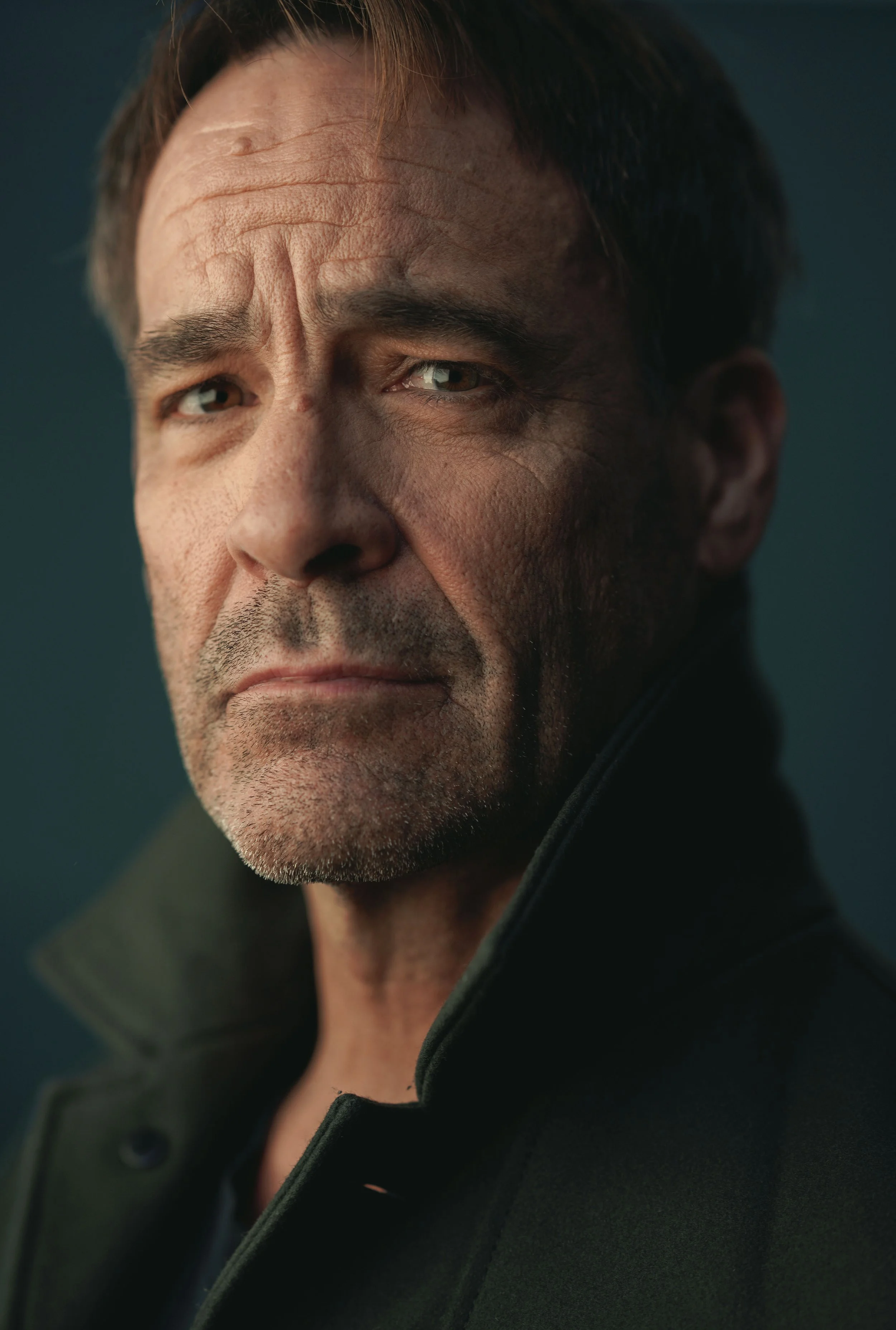 Close-up of a middle-aged man with short dark hair and a serious expression, wearing a black coat with a raised collar.