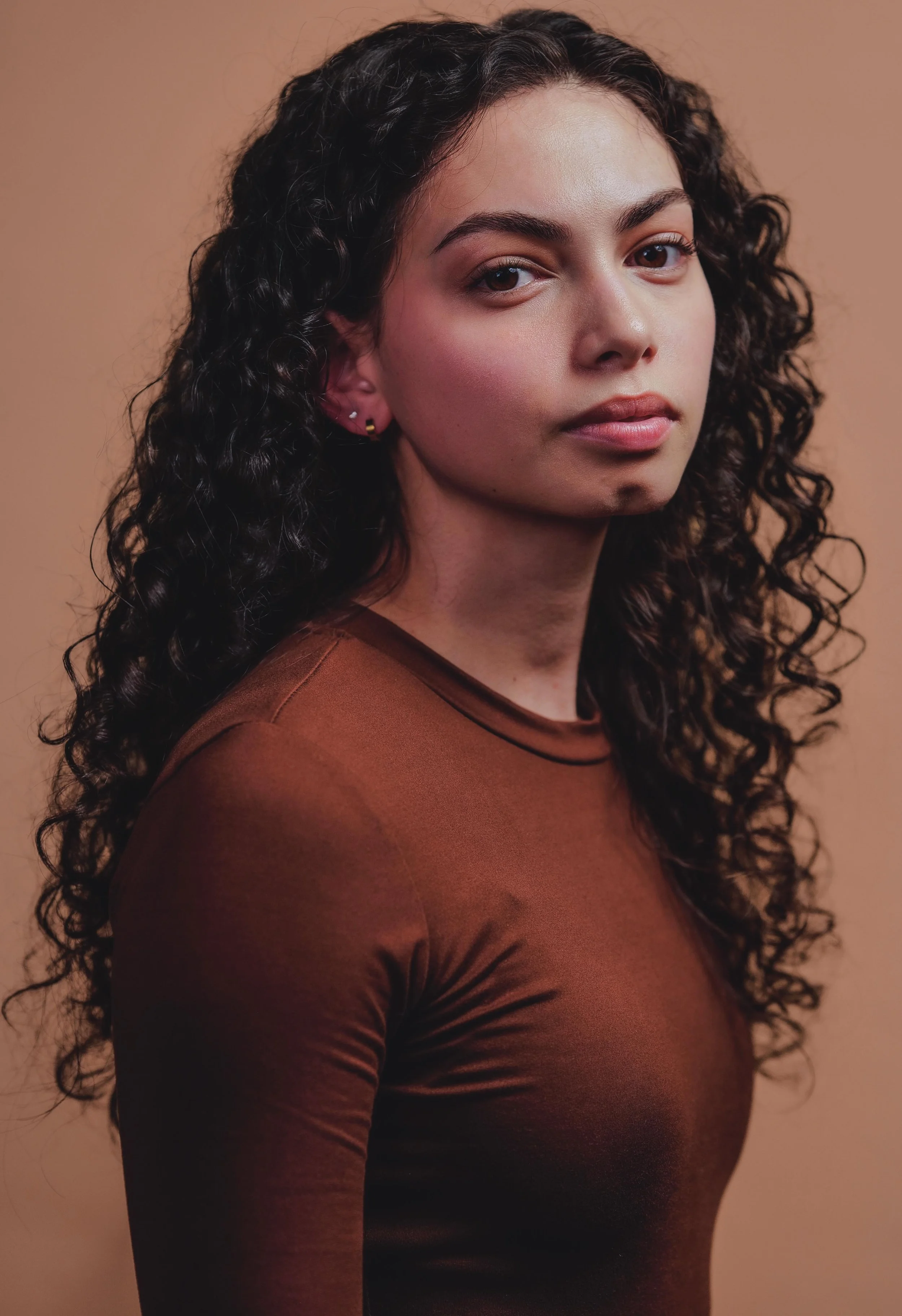 A young woman with dark, curly hair and light skin, wearing a brown long-sleeve top, looking at the camera with a neutral expression, standing against a beige background.