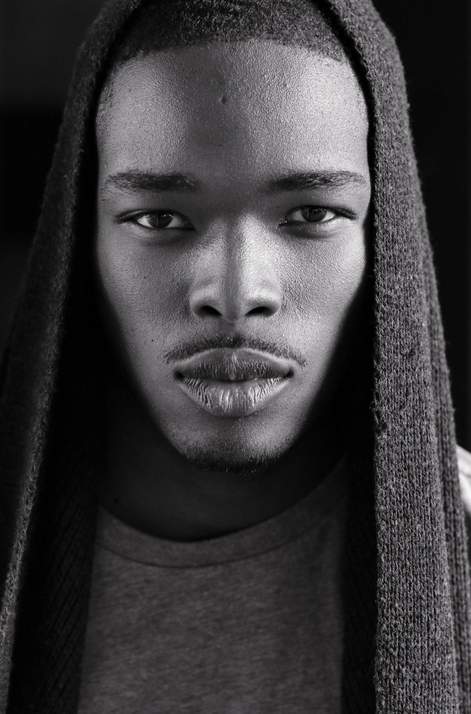 Close-up black-and-white portrait of a young man with dreadlocks, wearing a t-shirt, looking directly at the camera with a serious expression.