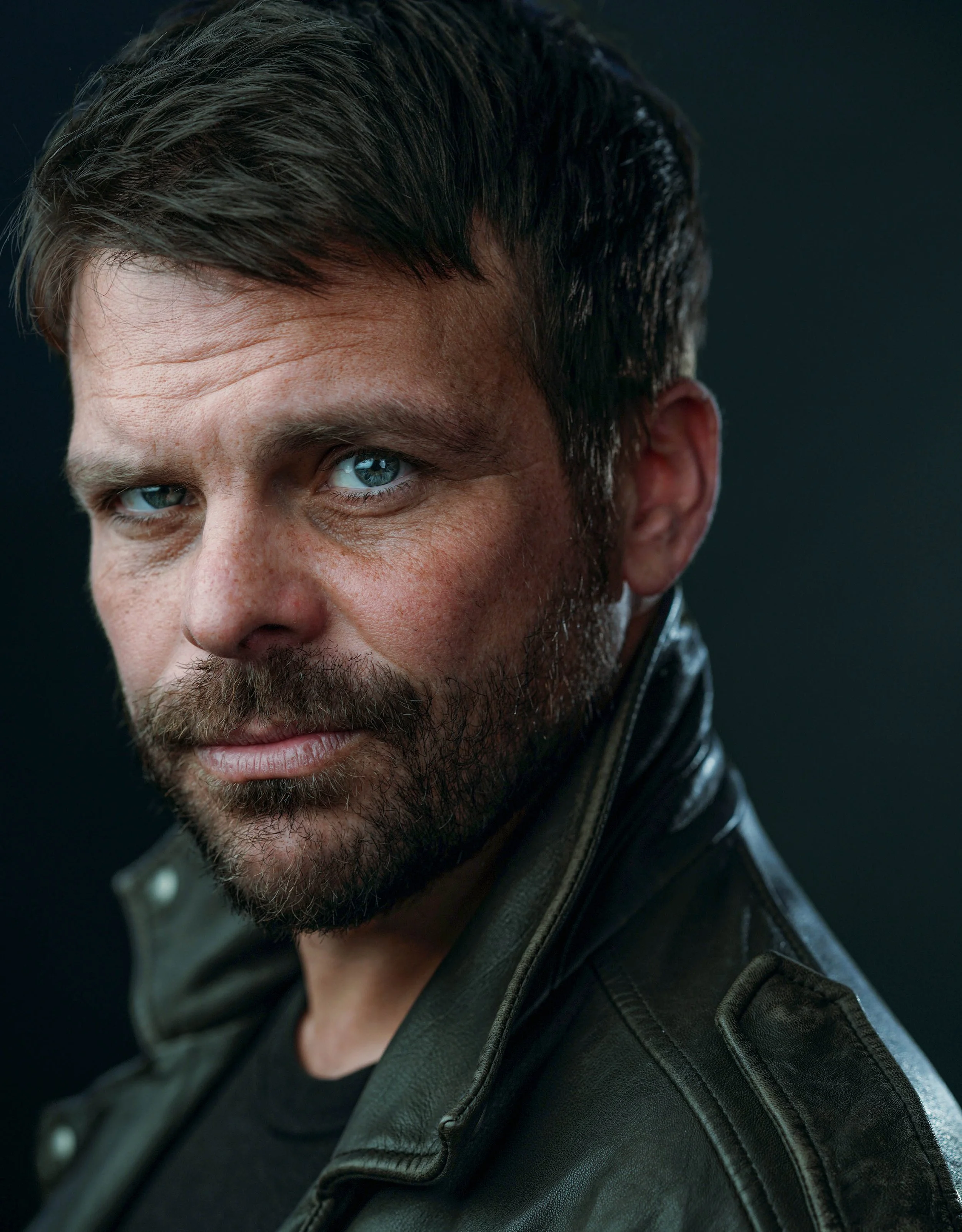 Close-up of a man with short dark hair, blue eyes, and a beard, wearing a black leather jacket, against a dark background.