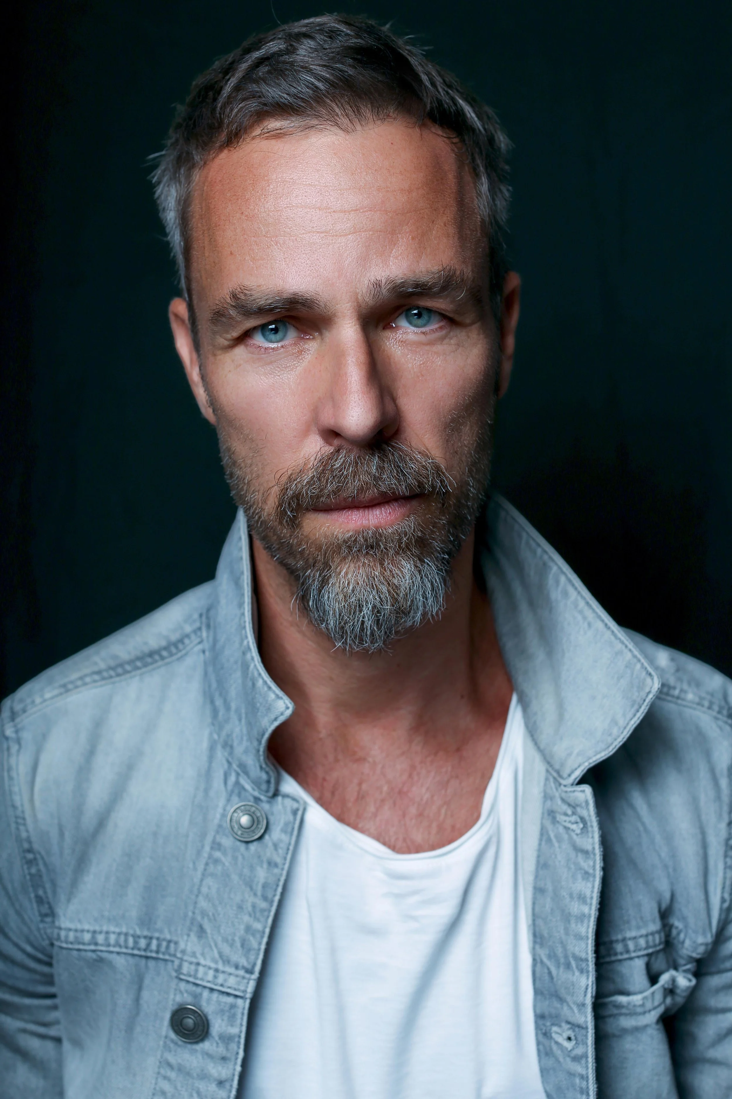 Close-up portrait of a middle-aged man with blue eyes, gray hair, and a beard, wearing a light denim jacket over a white t-shirt, against a dark background.