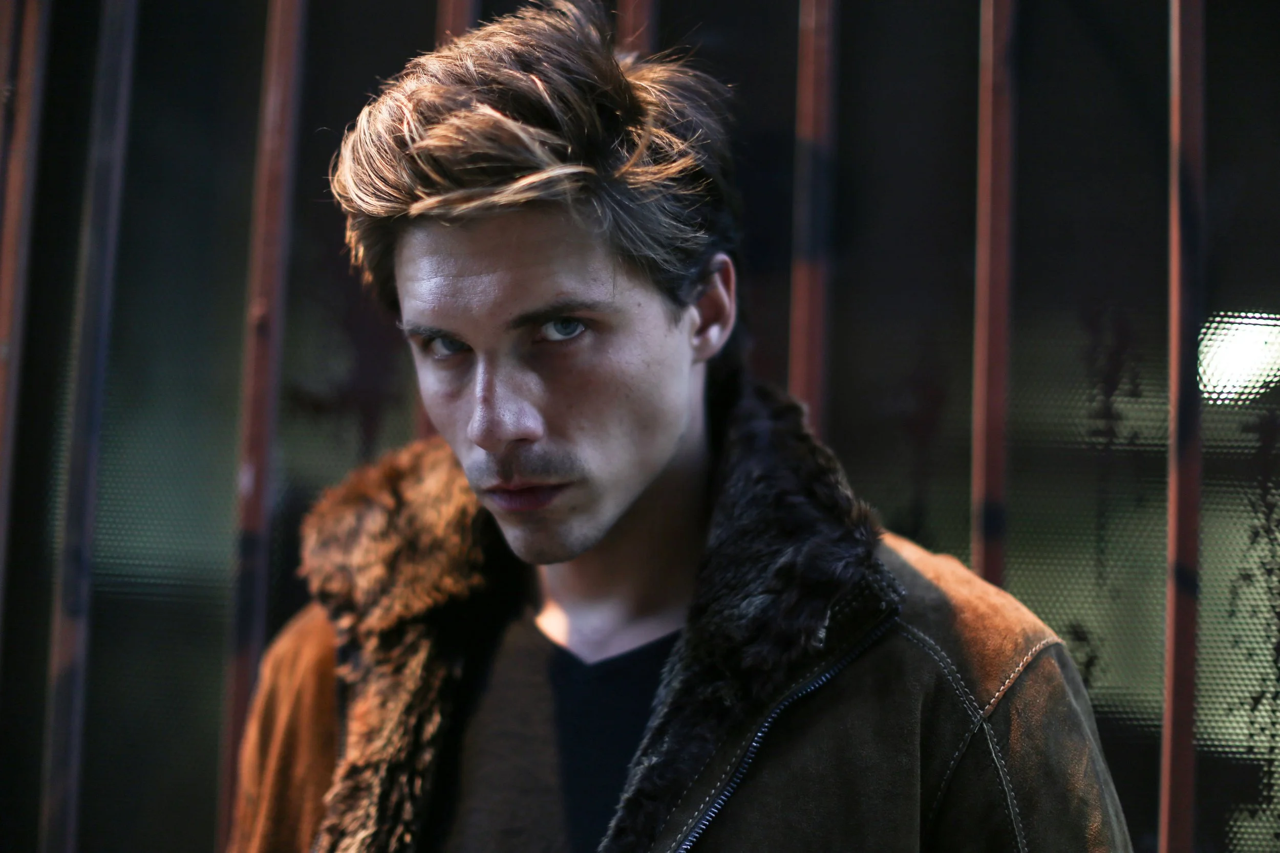A young man with styled hair and piercing blue eyes wearing a brown jacket with a fur collar, standing in front of a metal fence at night.