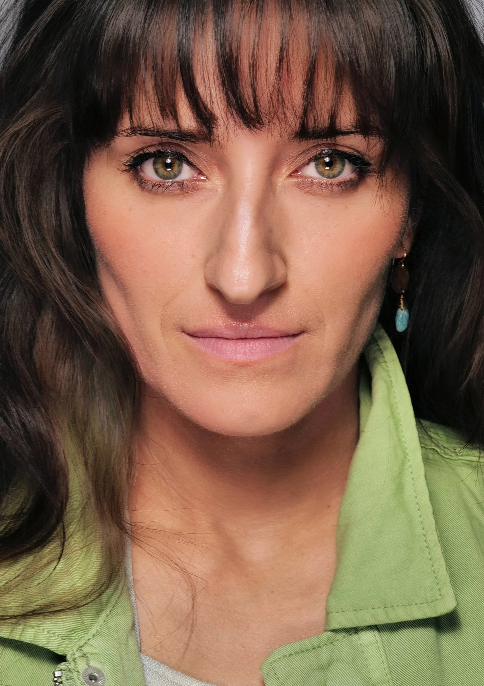 Close-up portrait of a woman with dark brown hair and bangs, green eyes, wearing turquoise earrings and a light green jacket.