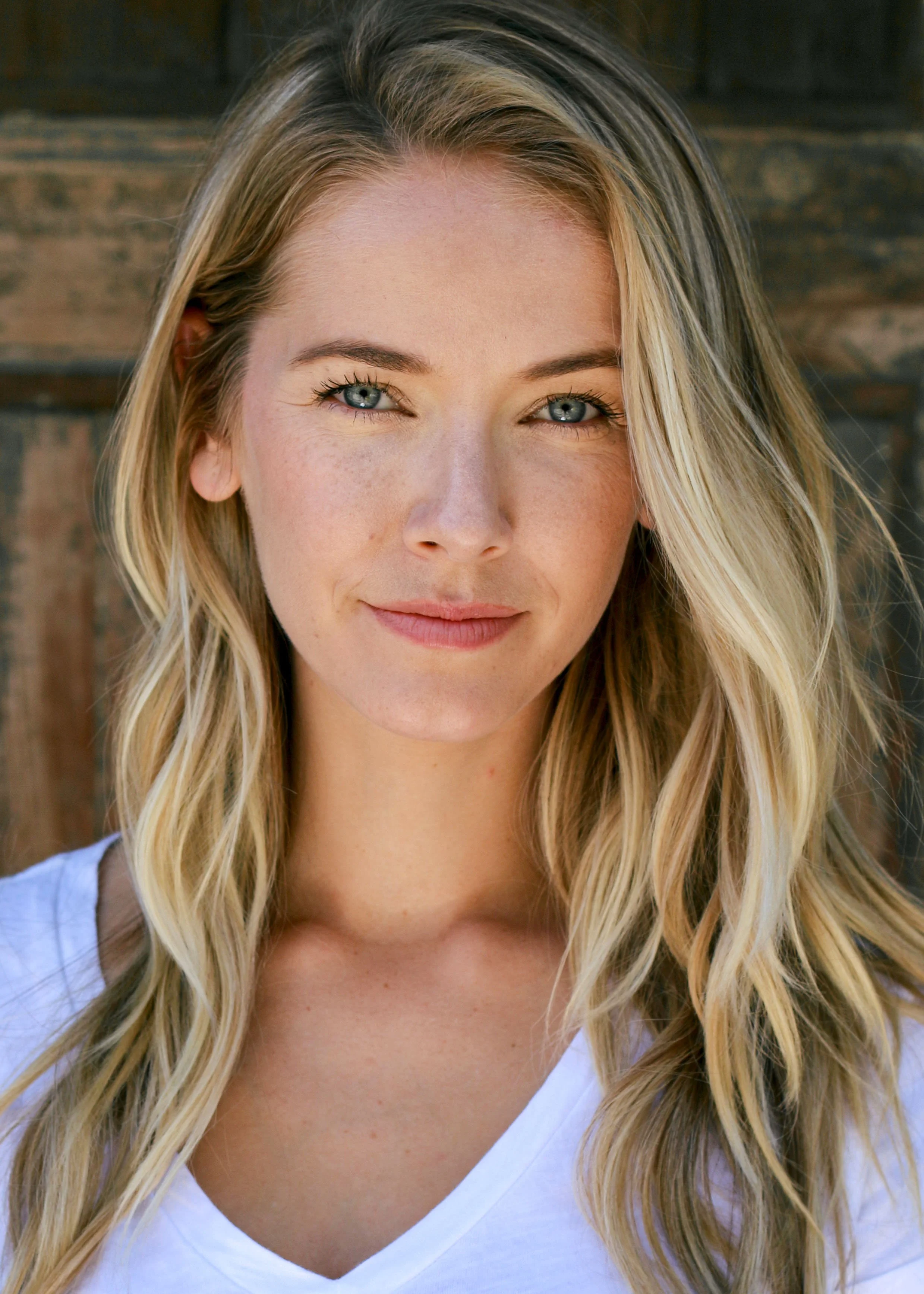 Close-up portrait of a young woman with blonde hair, blue eyes, and light skin, smiling lightly, wearing a white top, against a wooden background.