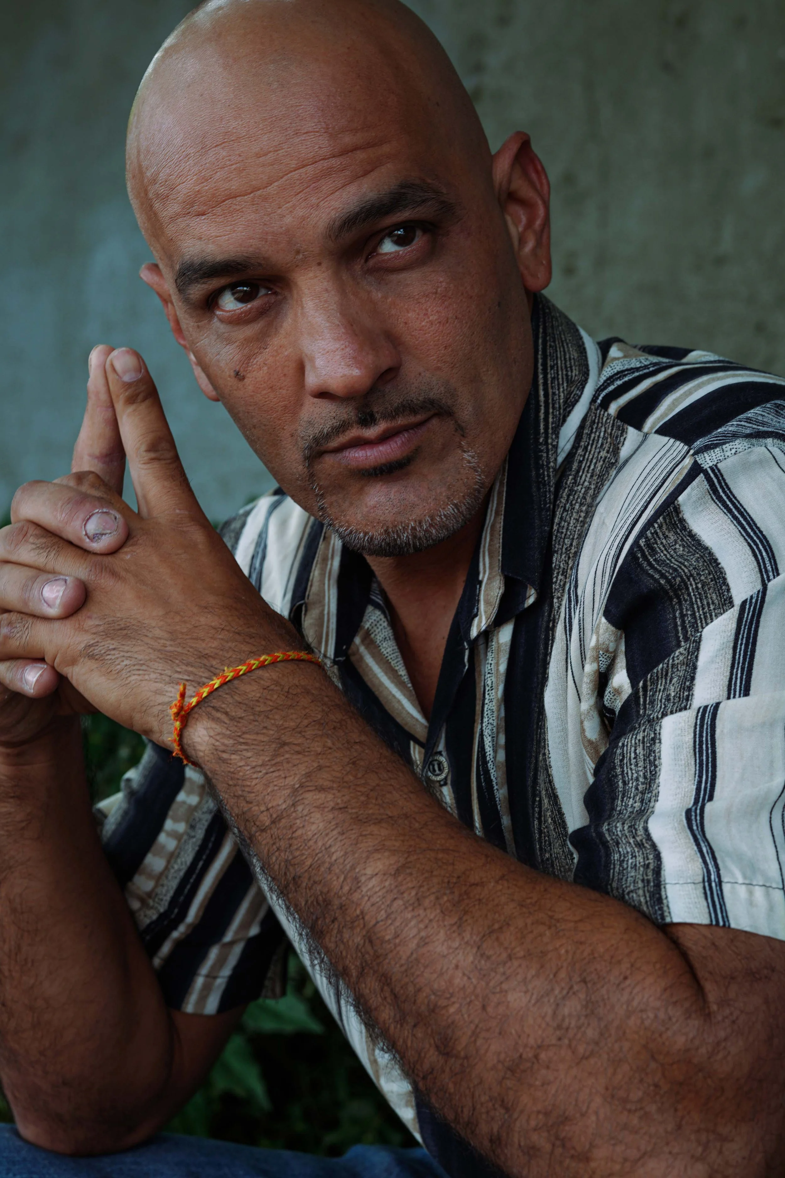 A close-up portrait of a bald man with a short beard, wearing a striped shirt, sitting outdoors with a serious expression, hands clasped, and a red and yellow bracelet on his wrist.