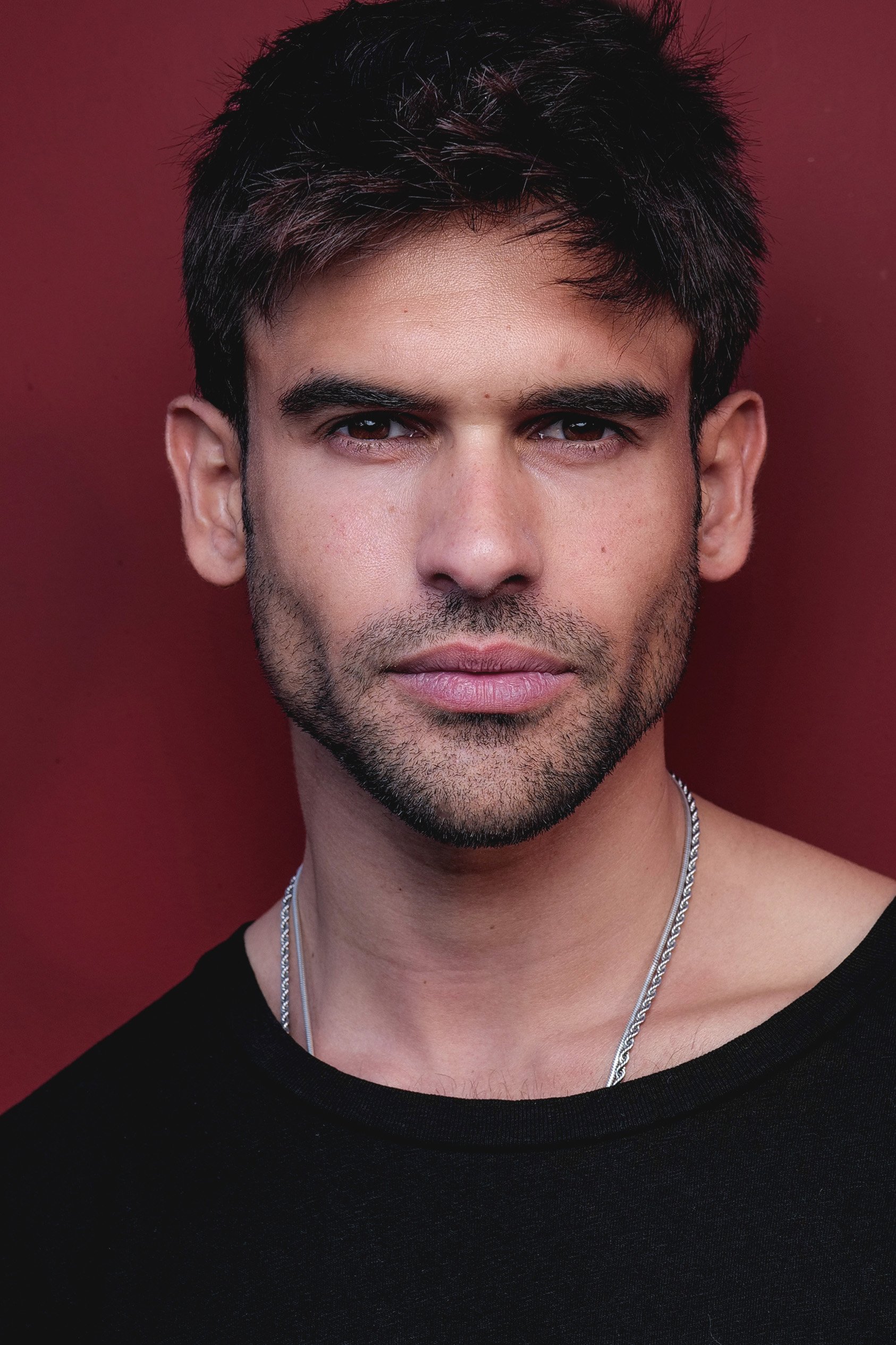 Close-up portrait of a young man with dark hair, wearing a black shirt and a silver chain necklace against a red background.