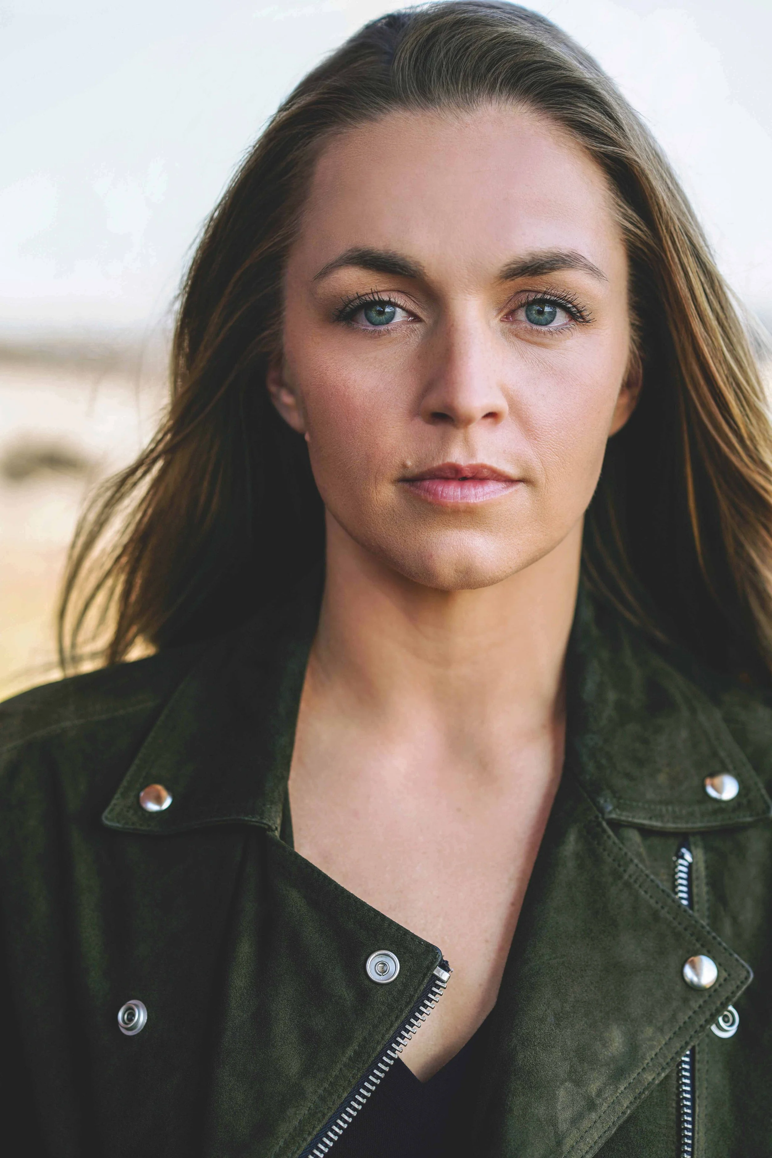 Close-up of a woman with long brown hair, fair skin, blue eyes, and natural makeup, wearing a dark green jacket.