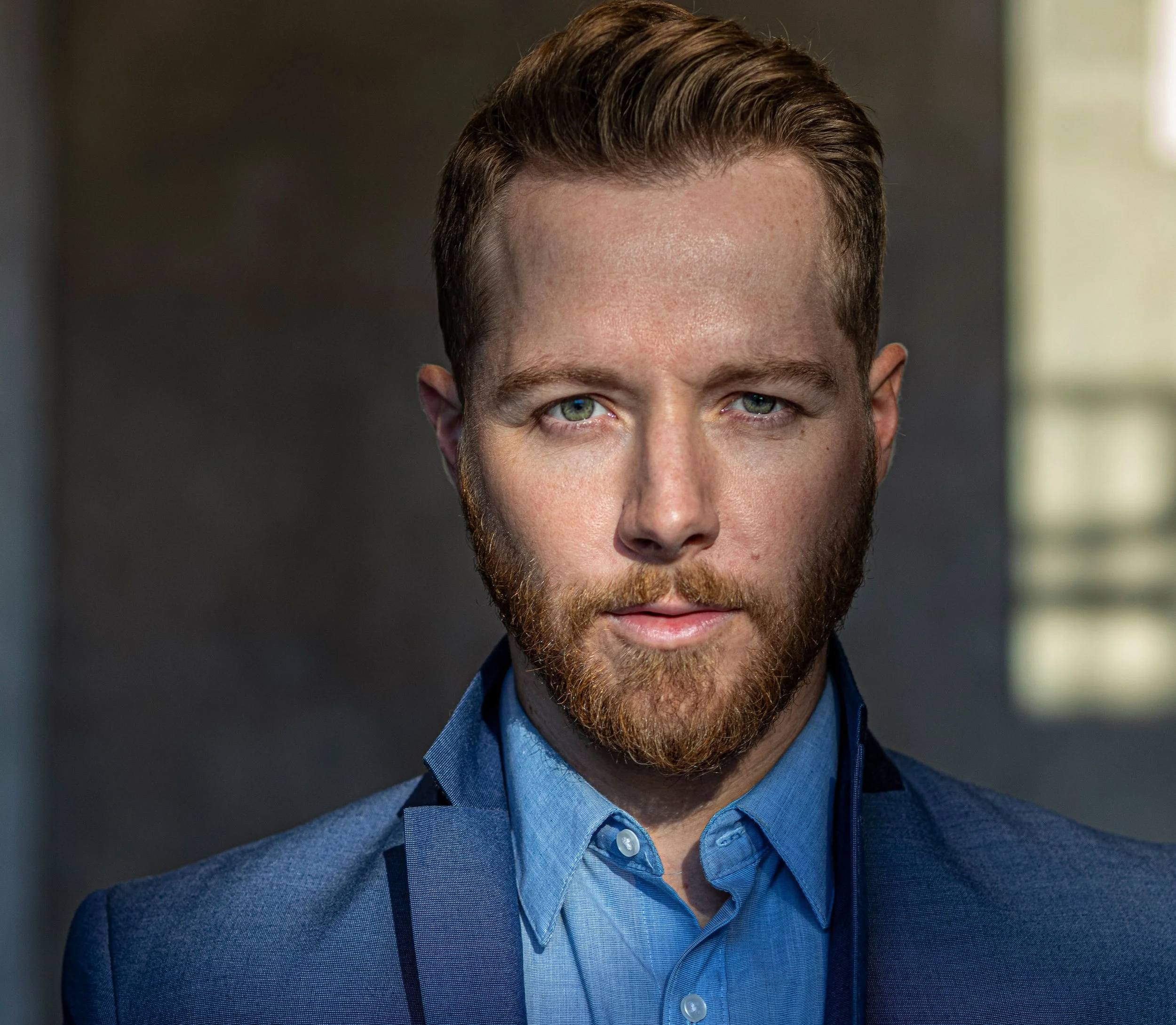 Close-up of a man with light brown hair, blue eyes, and a beard, wearing a blue dress shirt and a blue blazer, standing indoors with natural light coming through windows in the background.