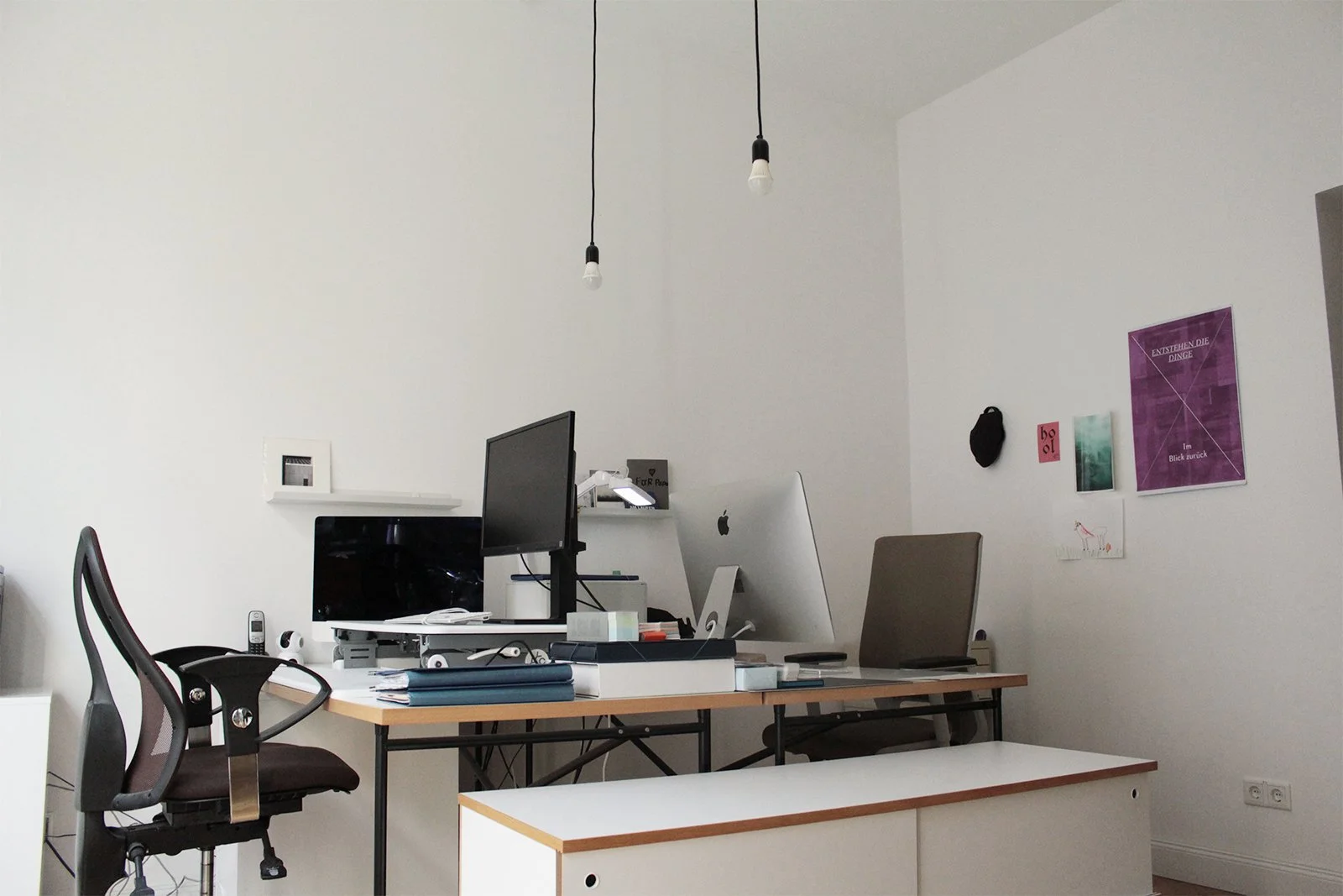 Modernes Büro mit zwei Schreibtischen, Monitoren, Laptops, ergonomischem Stuhl, hängenden Glühbirnen, weißen Wänden und dekorativen Bildern.