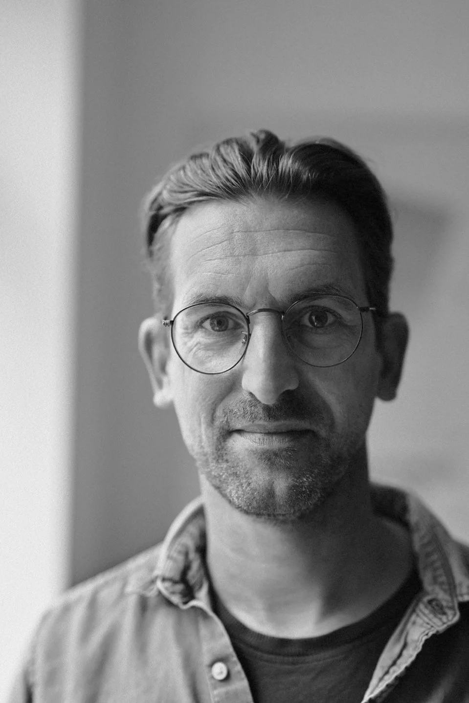 Schwarz-weiß-Foto eines Mannes mit Brille, der direkt in die Kamera schaut. Er trägt ein Hemd und hat kurzes, nach hinten gekämmtes Haar.