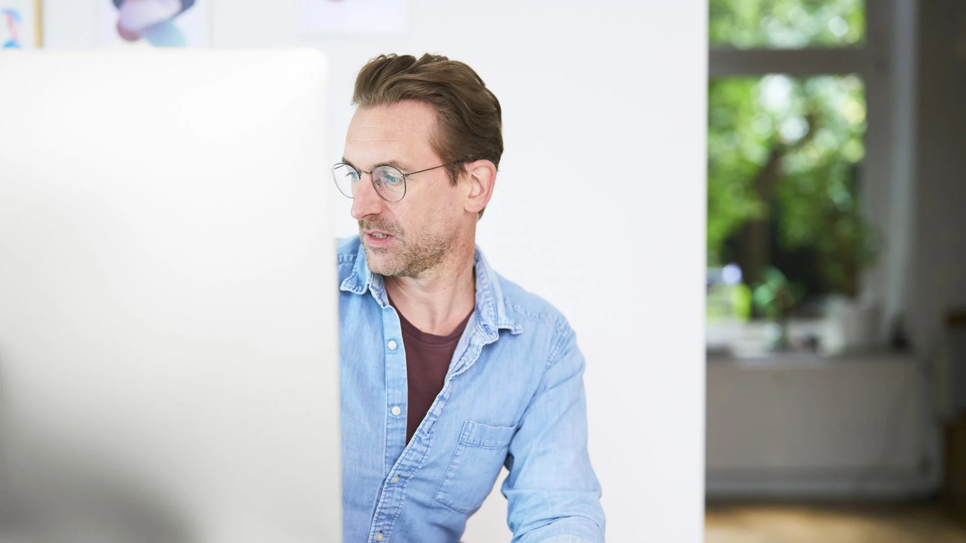 Mann mit Brille in einem hellen Raum vor einem Computer sitzend, schaut zur Seite, trägt ein blaues Hemd.