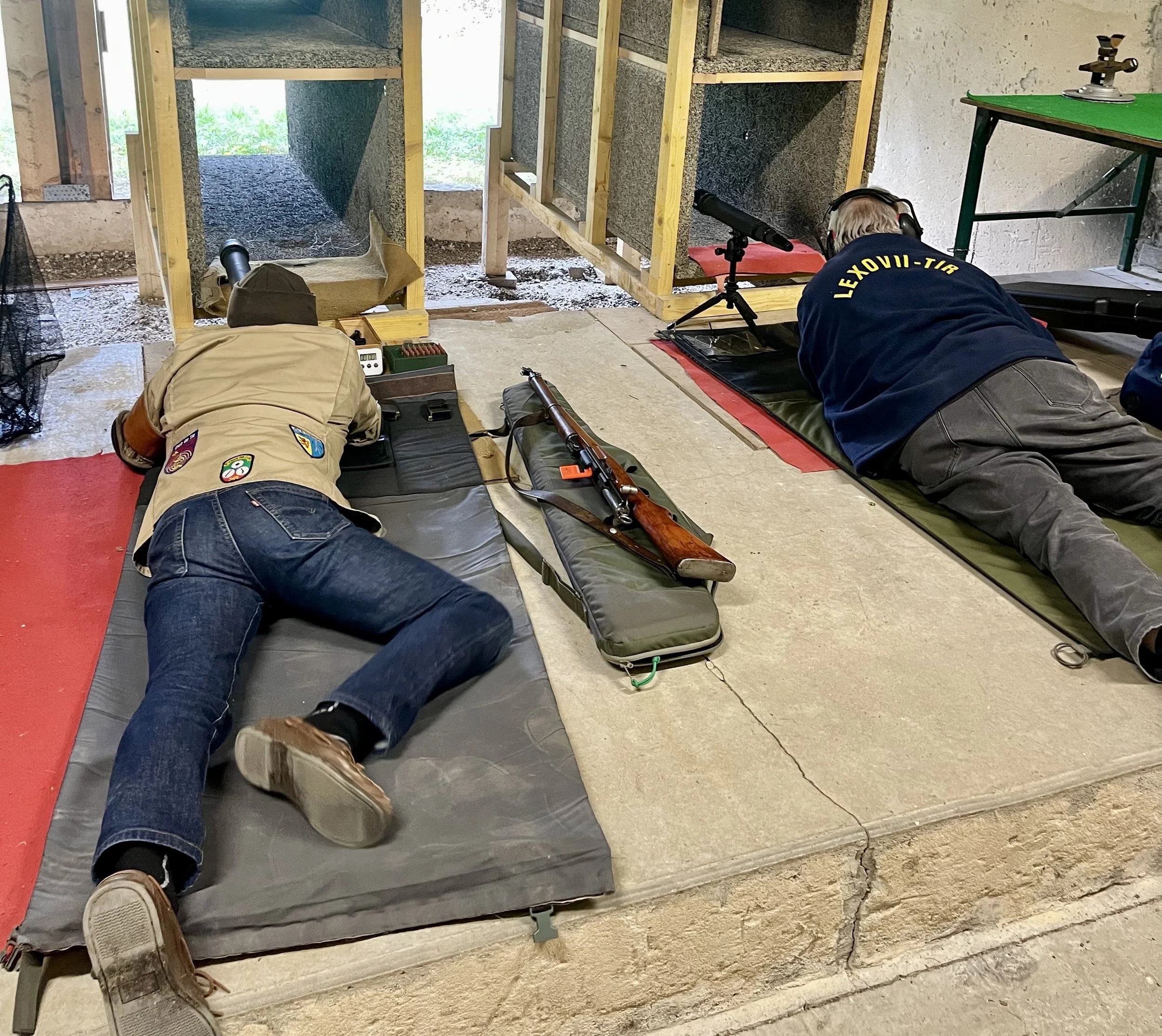 Homme avec casque de protection et lunettes, manipulant un fusil avec lunette de visée, allongé sur le sol.